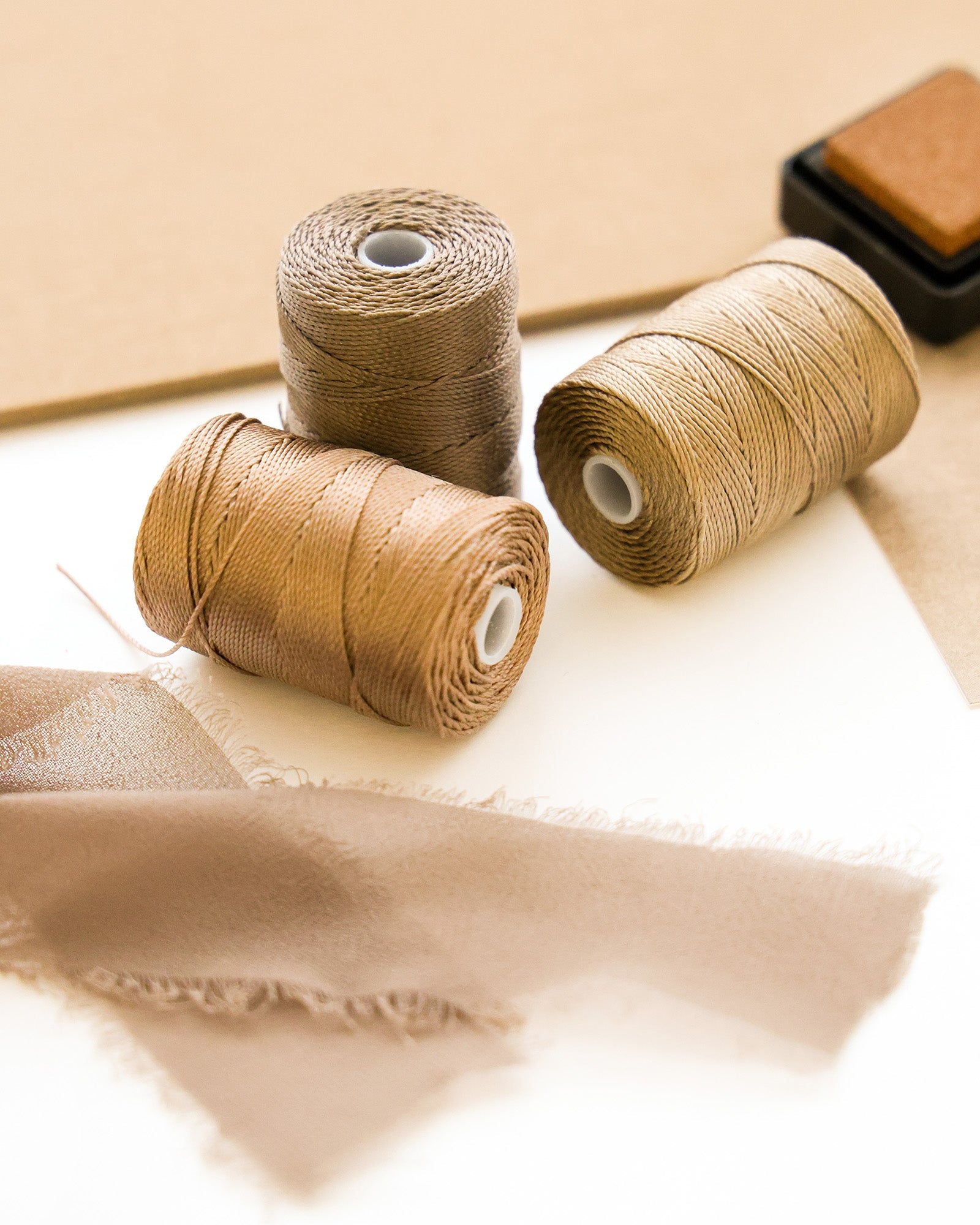 Drei Spulen mit hellbraunem und braunem Garn mit Stoffmustern und einem braunen Stempelkissen auf einer weißen Fläche.