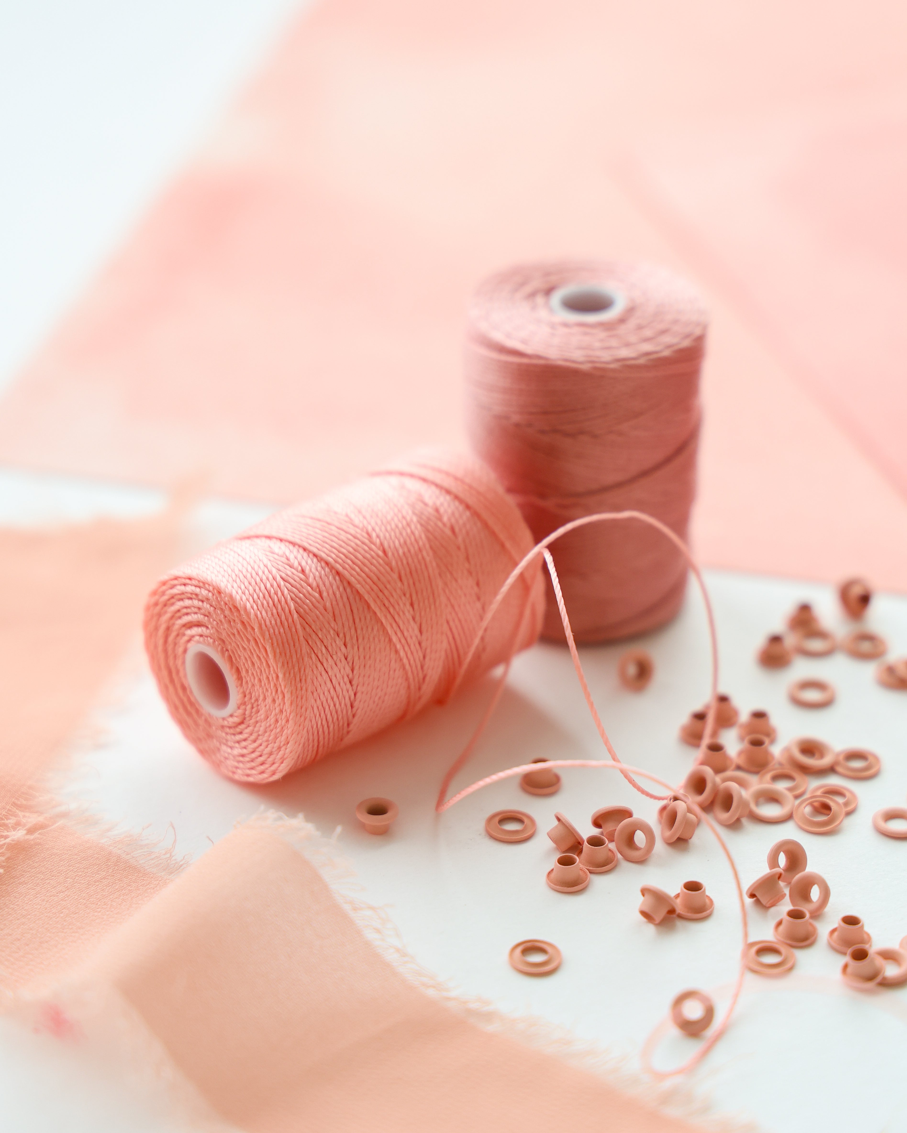 Zwei Spulen mit rosa Faden und verstreuten rosa Perlen auf einer weißen Fläche mit rosa Stoffstücken.