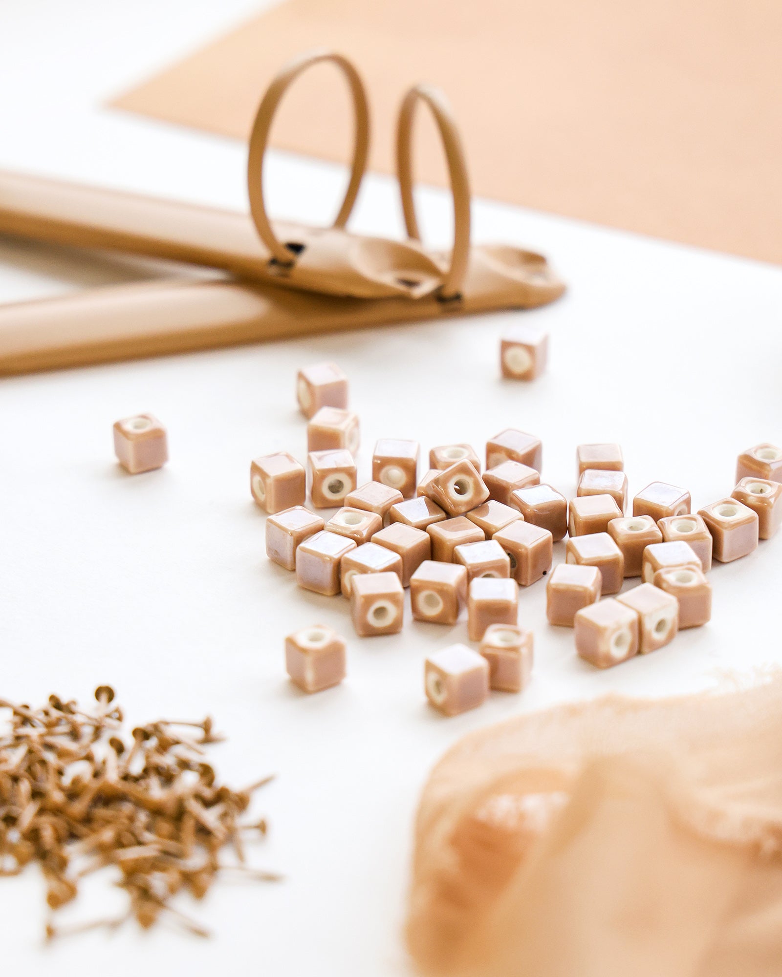 Ein Stapel beigefarbener quadratischer Perlen, Scheren, Nadeln und Spitzen auf einer weißen Fläche mit hellbraunen Akzenten.