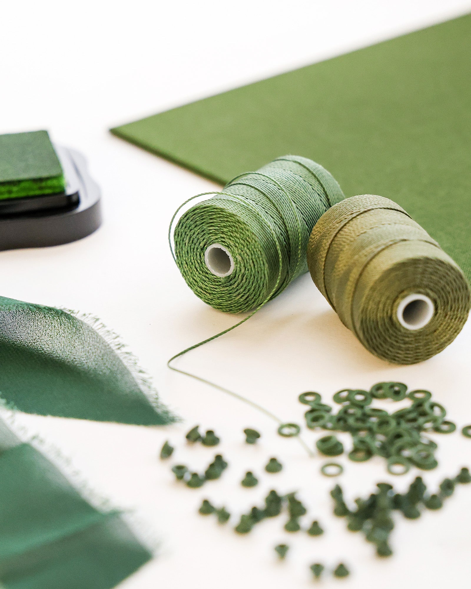 Grüne Garnspulen, Stoff, Knöpfe und Stempelkissen auf einer weißen Fläche angeordnet.