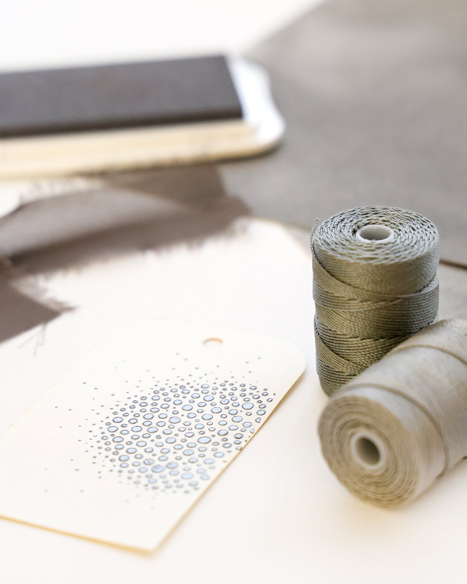 Graue Garnspulen und eine gemusterte Karte auf einer weißen Fläche mit Stoff und einem Notizbuch im Hintergrund.