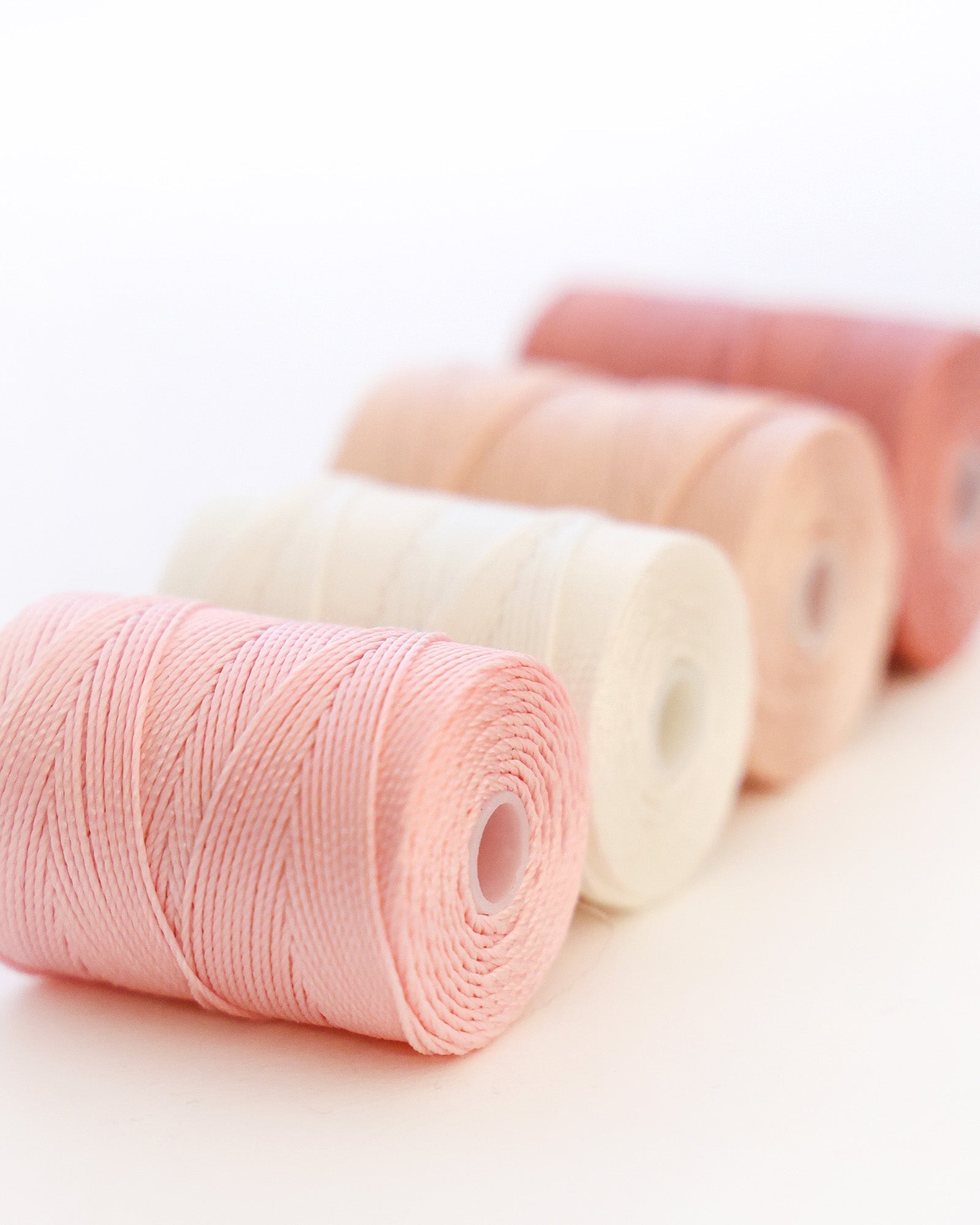Vier Garnspulen in Pastellrosa-, Creme- und Pfirsichtönen sind in einer diagonalen Linie angeordnet.