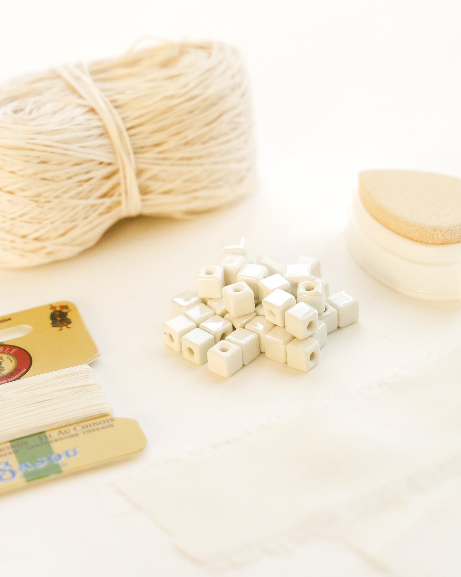 Ein Bündel Schnur, weiße quadratische Perlen, Fadenkarten und ein kleines rundes Schaumstoffstück auf einer weißen Fläche.