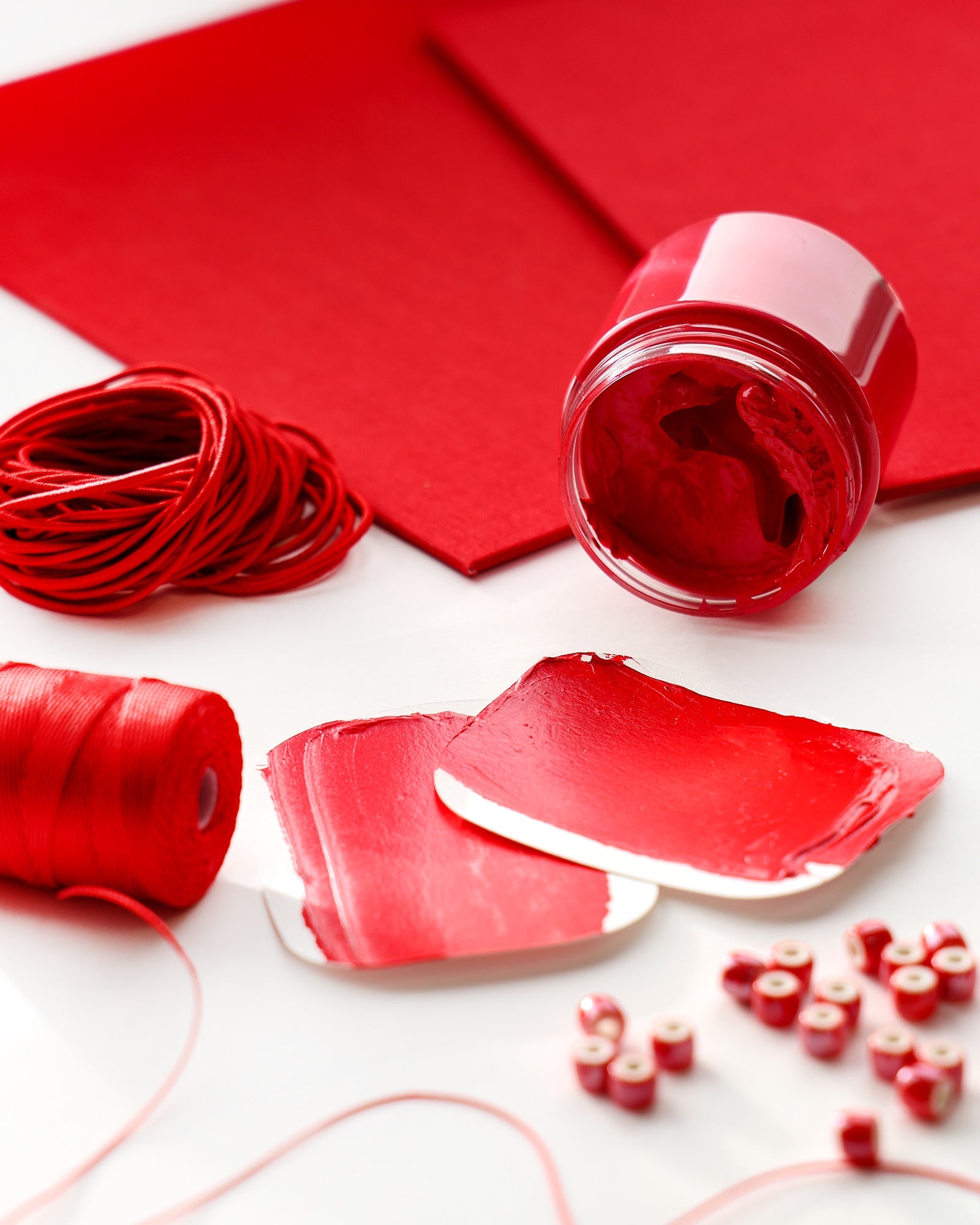 Rote Farbe, Faden, Papier und Perlen, die auf einer weißen Fläche angeordnet sind, wobei die Farbe mit Spachteln verteilt wird.