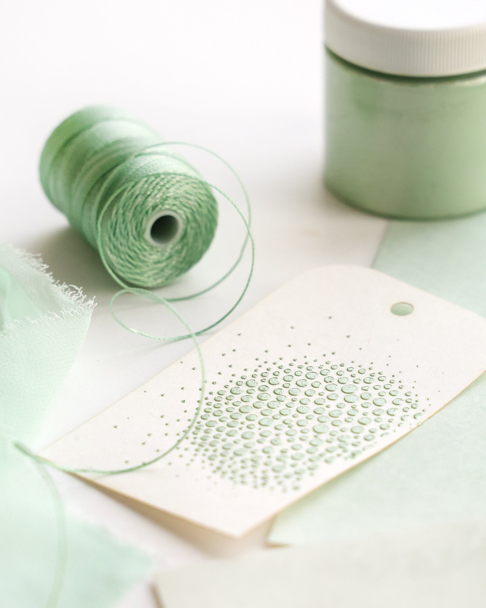 Mintgrüner Faden, ein Glas und Stoff neben einer Karte mit mintgrüner gepunkteter Grafik auf einer weißen Fläche.