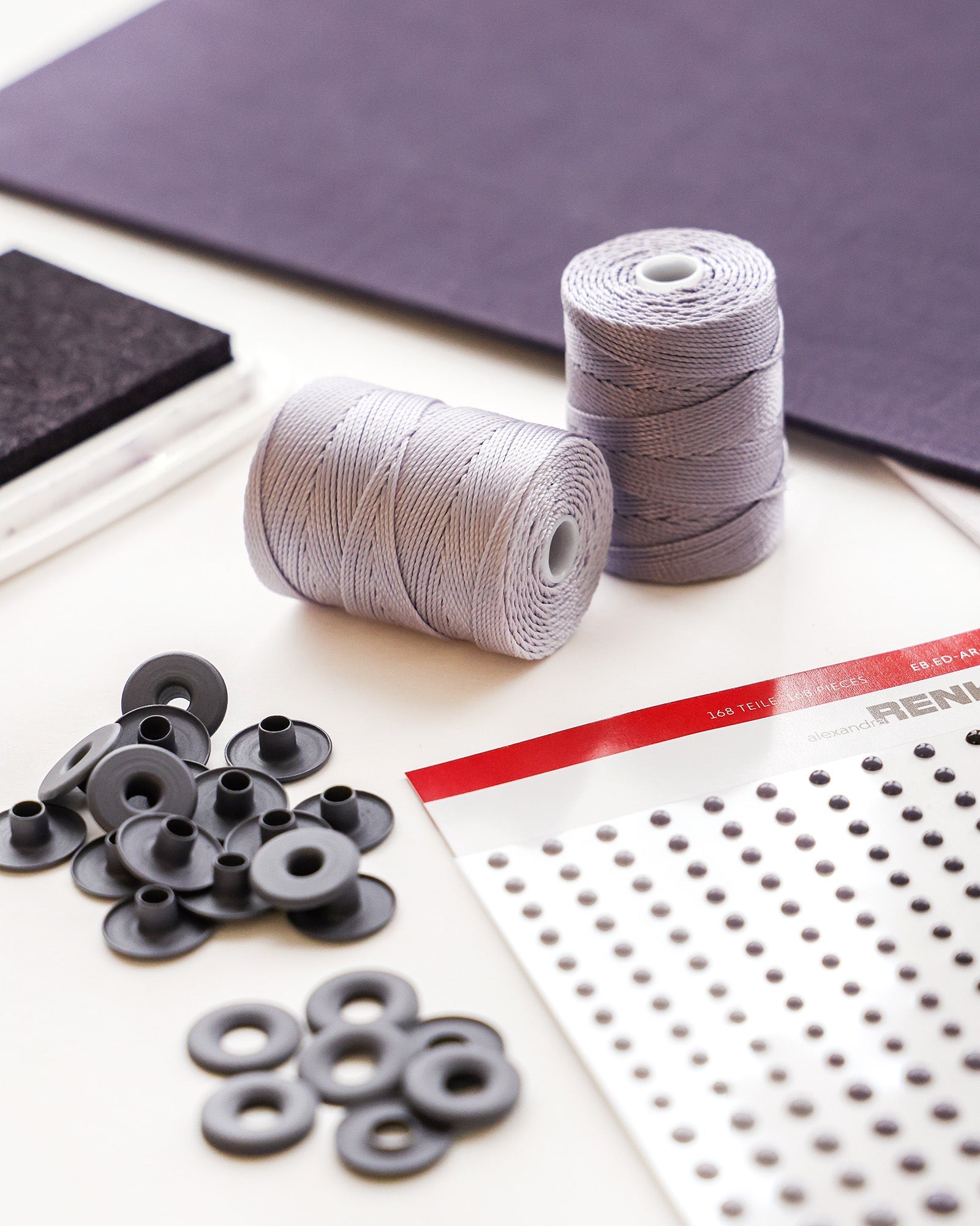 Zwei Spulen mit grauem Garn, schwarze Ösen und ein Blatt mit Klebepunkten auf einem Tisch.