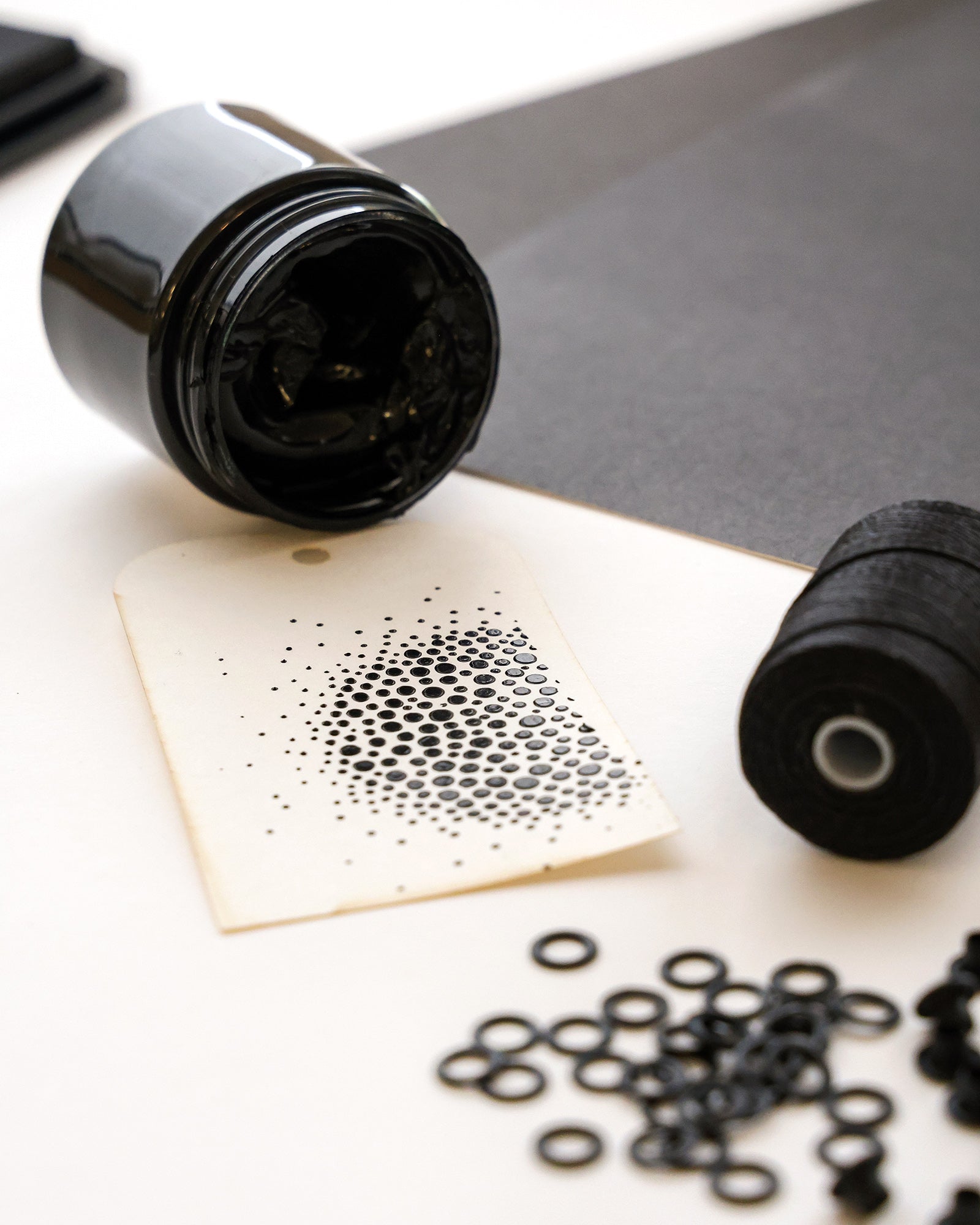 Ein Glas mit schwarzer Tinte, ein schwarzer Faden und Ringe neben einem Papier mit einem Punktmuster auf einer weißen Fläche.