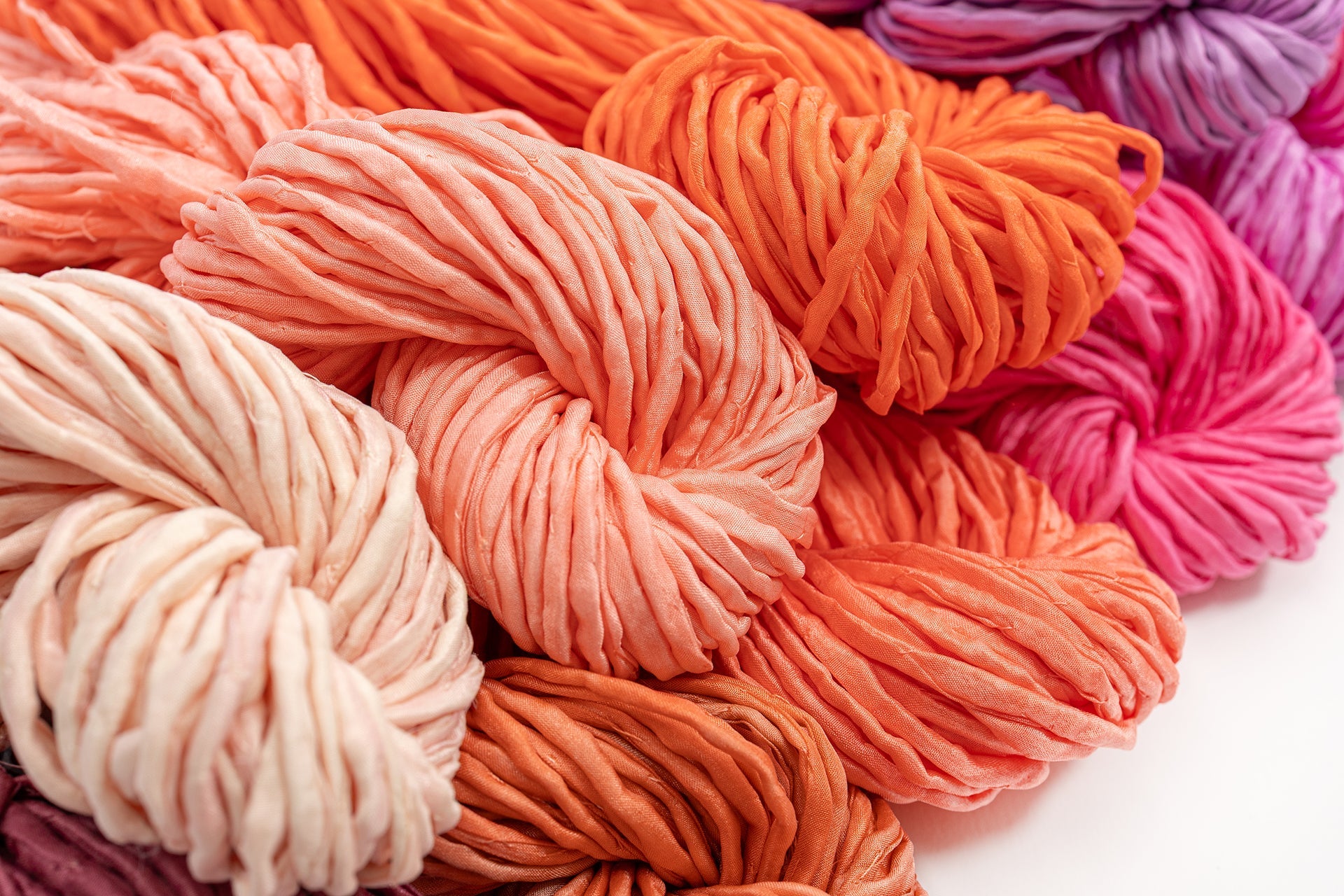 Mehrere Garnstränge in Rosa-, Orange-, Pfirsich- und Violetttönen sind dicht nebeneinander angeordnet und bilden so ein farbenfrohes und strukturiertes Muster.