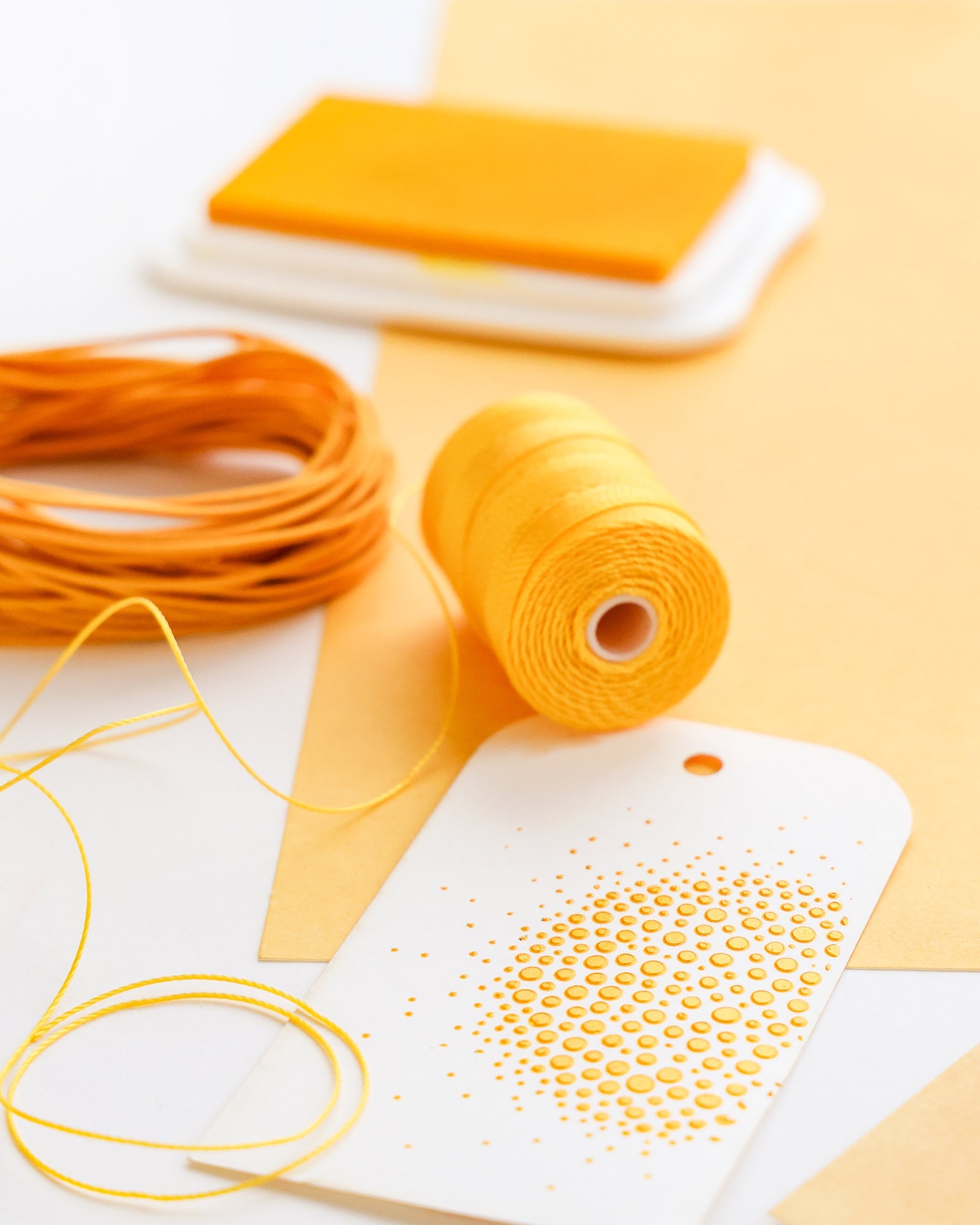 Gelber Faden, Kordel und Stempelkissen neben einem weißen Geschenkanhänger mit gelbem Punktmuster auf gelbem Papierhintergrund.