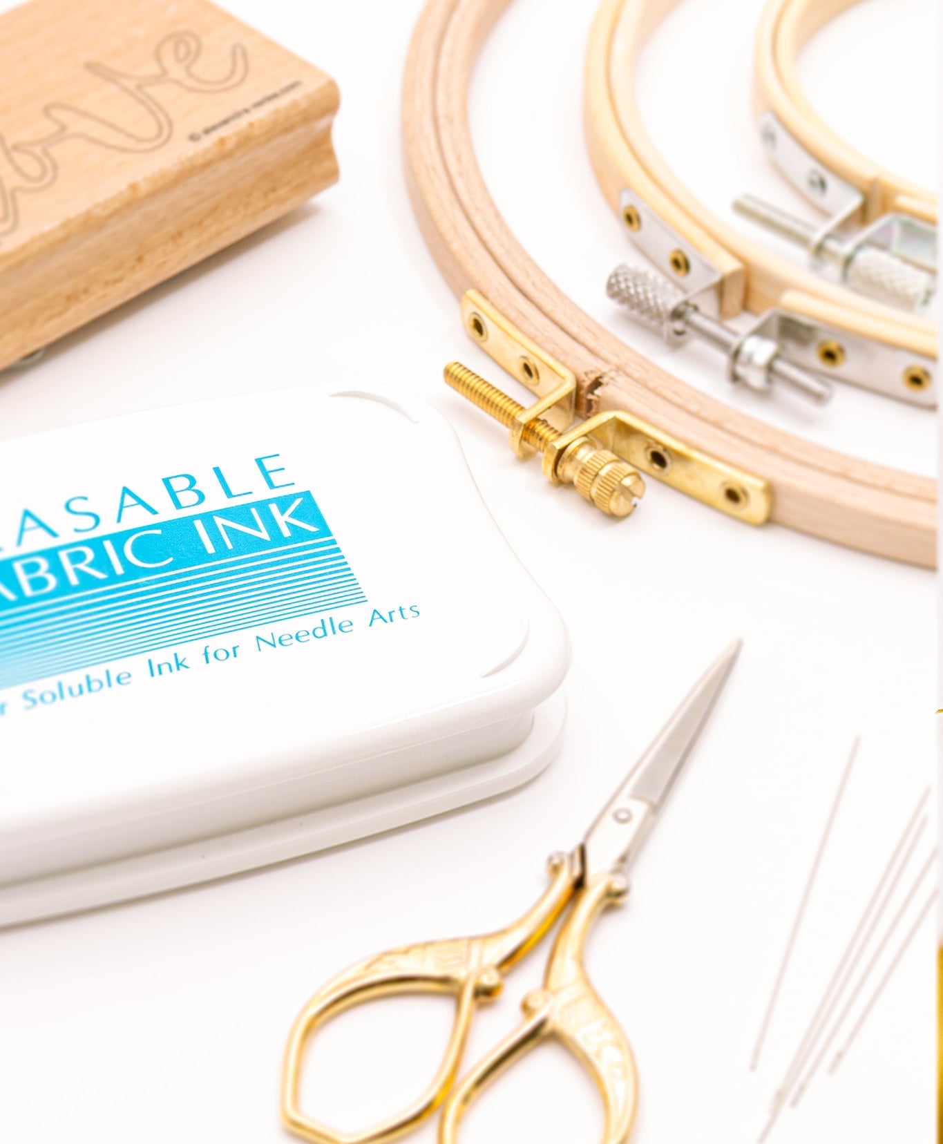 Stickutensilien auf einem weißen Untergrund, darunter Stickrahmen, eine goldene Schere, Nadeln, eine Schachtel mit der Aufschrift „Erasable Fabric Ink“ und ein Teil eines Holzblocks.