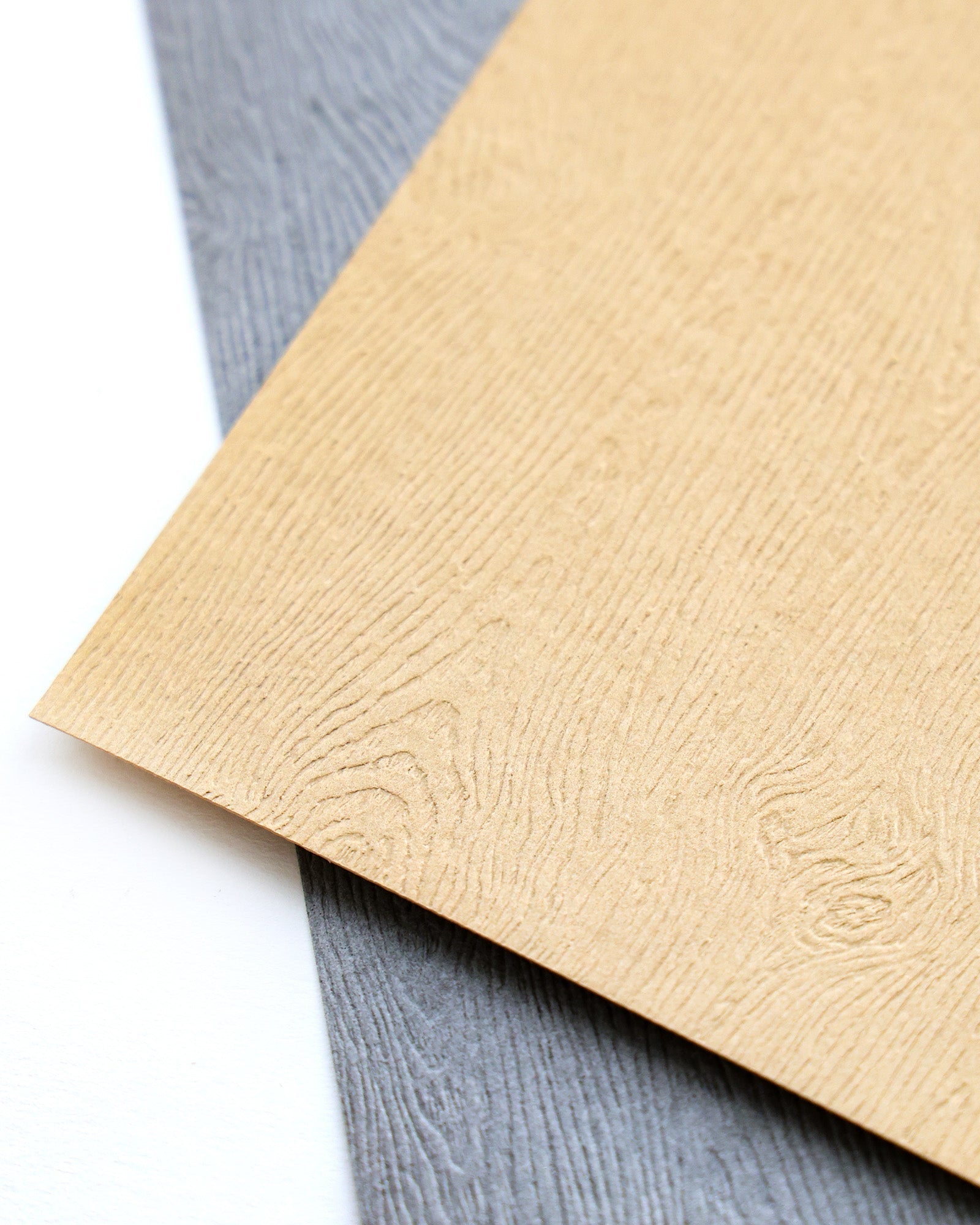 Zwei Blätter aus strukturiertem Papier, ein hellbraunes und ein graues, überlappen sich auf einer weißen Fläche.