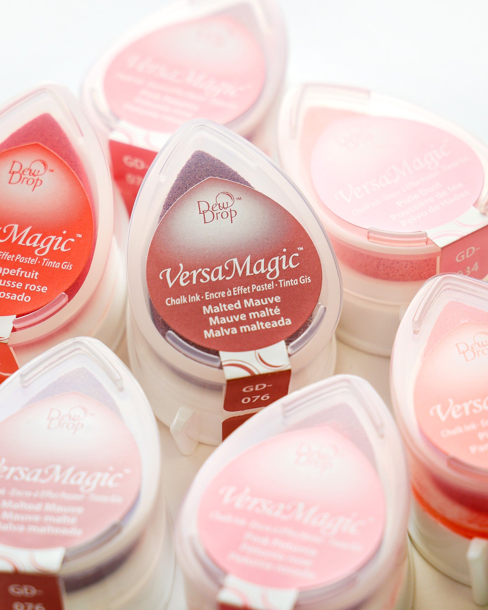 Mehrere VersaMagic-Tintenkissen in tropfenförmigen Behältern, meist in Rosa- und Lila-Tönen, sind aneinander gereiht.