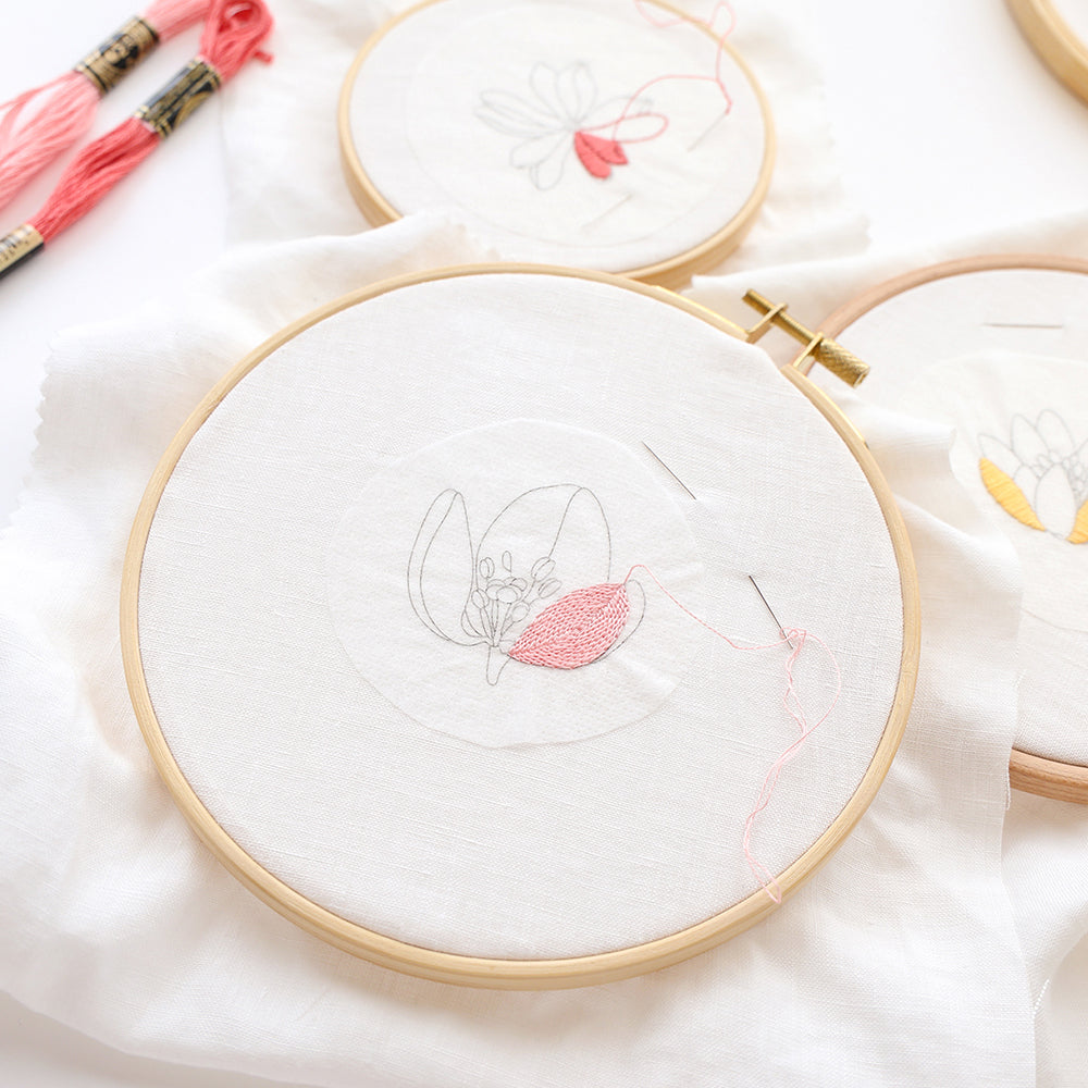 Ein Stickrahmen mit einem Blumenmotiv in Arbeit, rosa Garn und Nadel auf weißem Stoff von Alexandra Renke – ideal für kreative Projekte oder zum Üben deiner Lieblings-Stickvorlagen.