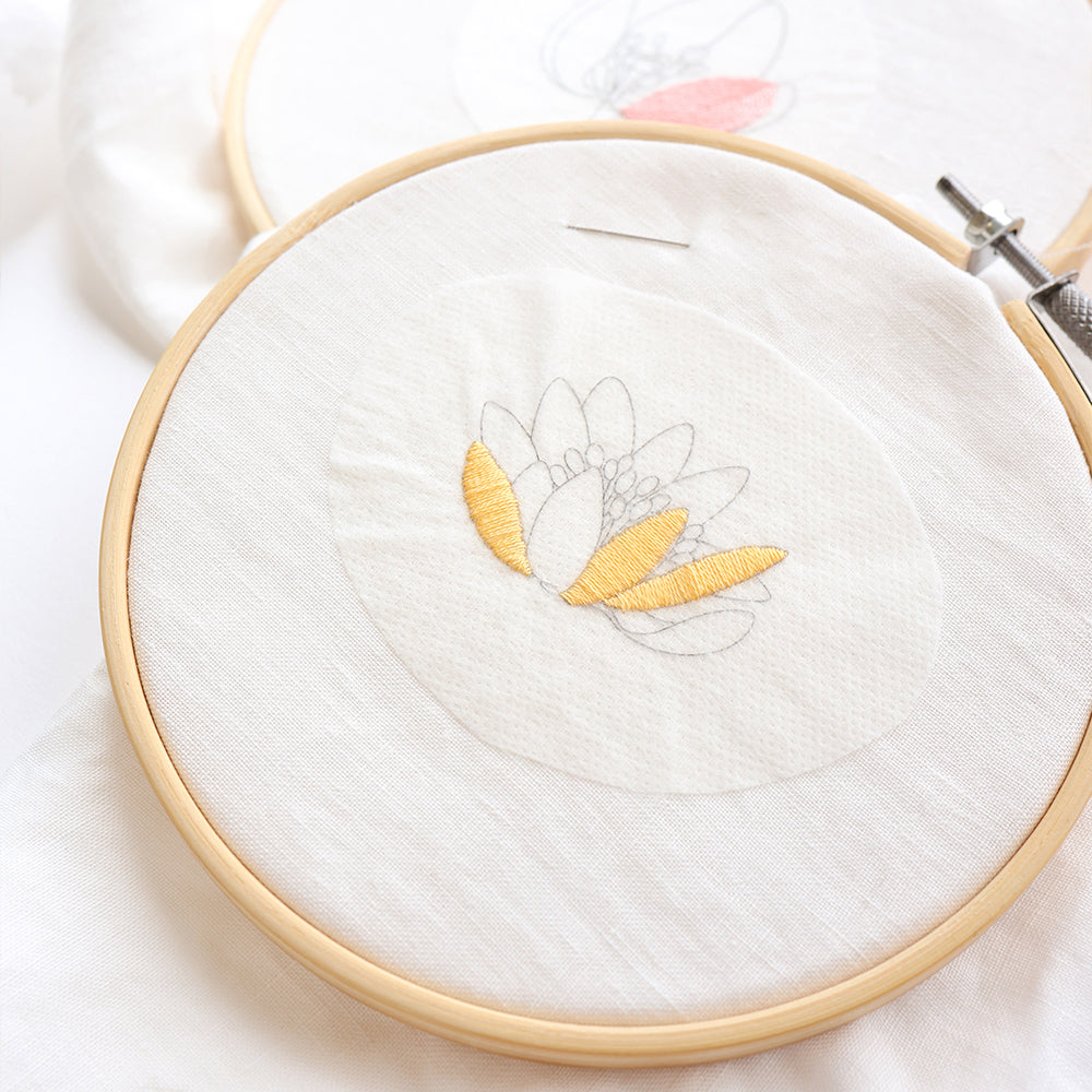 Stickrahmen mit einem Blumenumriss von Alexandra Renke; einige Blütenblätter sind mit gelbem Garn auf weißem Stoff gestickt – eine reizvolle Idee für Stickprojekte oder bei der Verwendung einer selbstklebenden Stickvorlage.