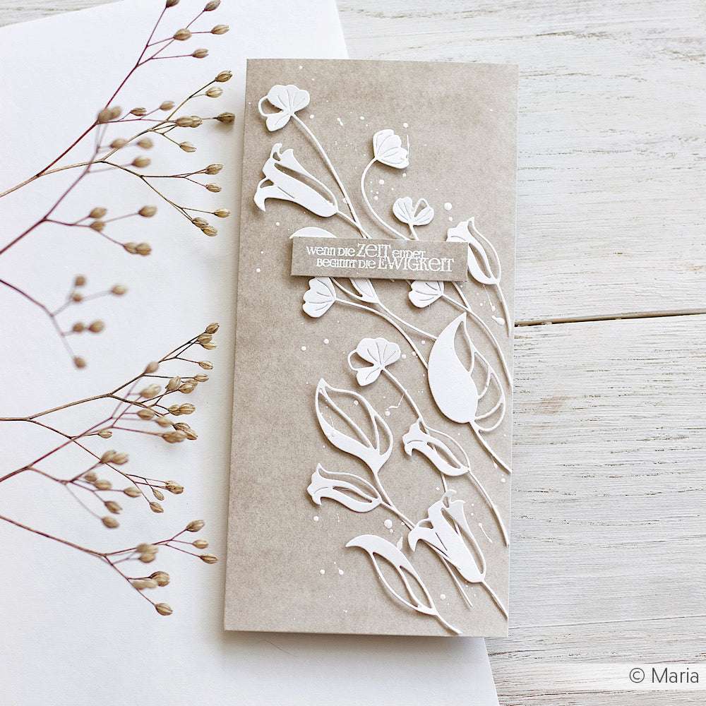 Eine handgefertigte Karte mit weiß geprägten Blumen auf Alexandra Renke Designpapier Mimi Schlamm, ausgestellt auf einer weißen Holzfläche - eine Präsentation kreativer Papierprojekte und Buchbinderei.