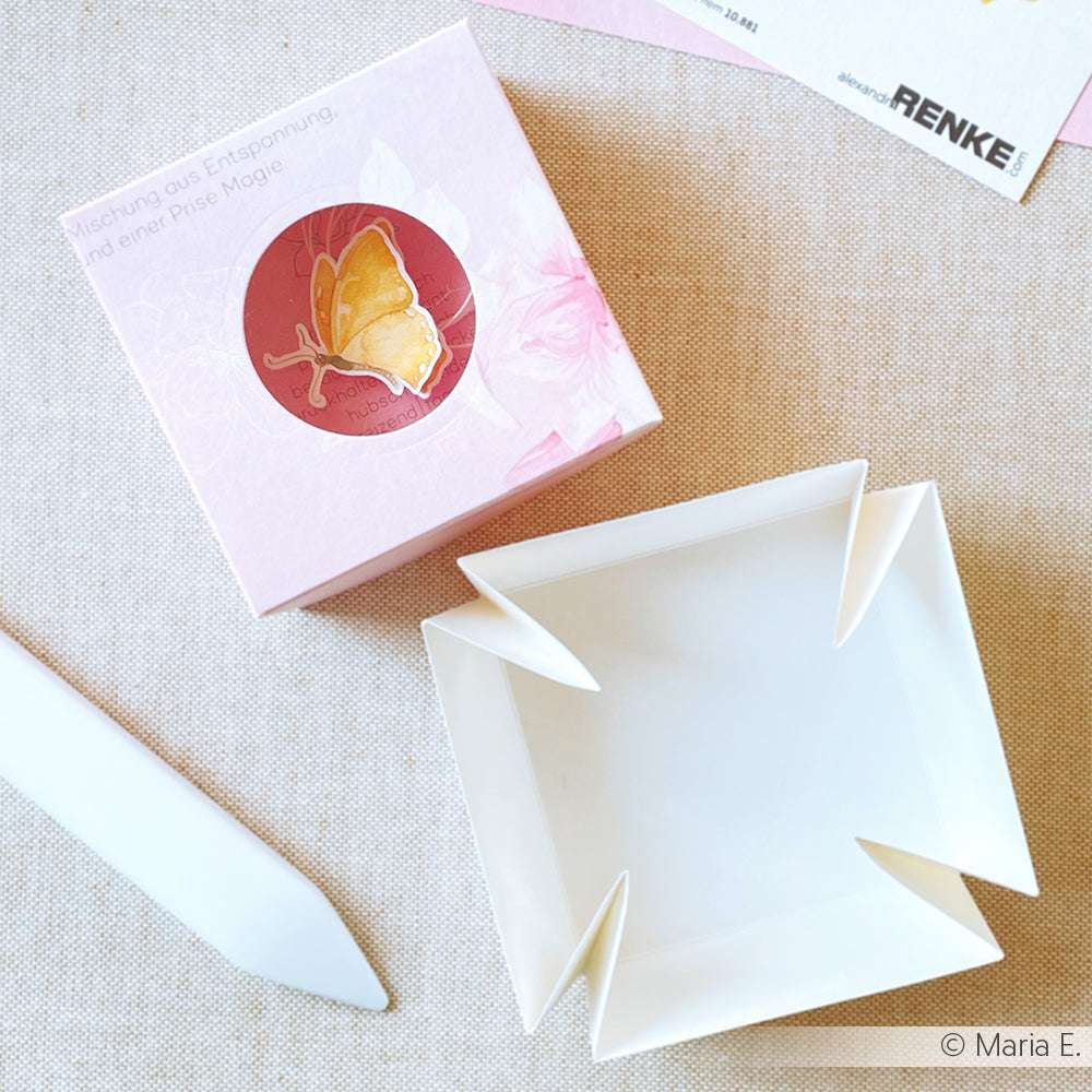 Rosa Schachtel aus Alexandra Renke Designpapier Schmetterlinge mit einem Schmetterlingsmotiv, neben einem aufgeklappten weißen Papierprojekte-Tablett und einem weißen Werkzeug, alles auf einer beigen Fläche angeordnet.