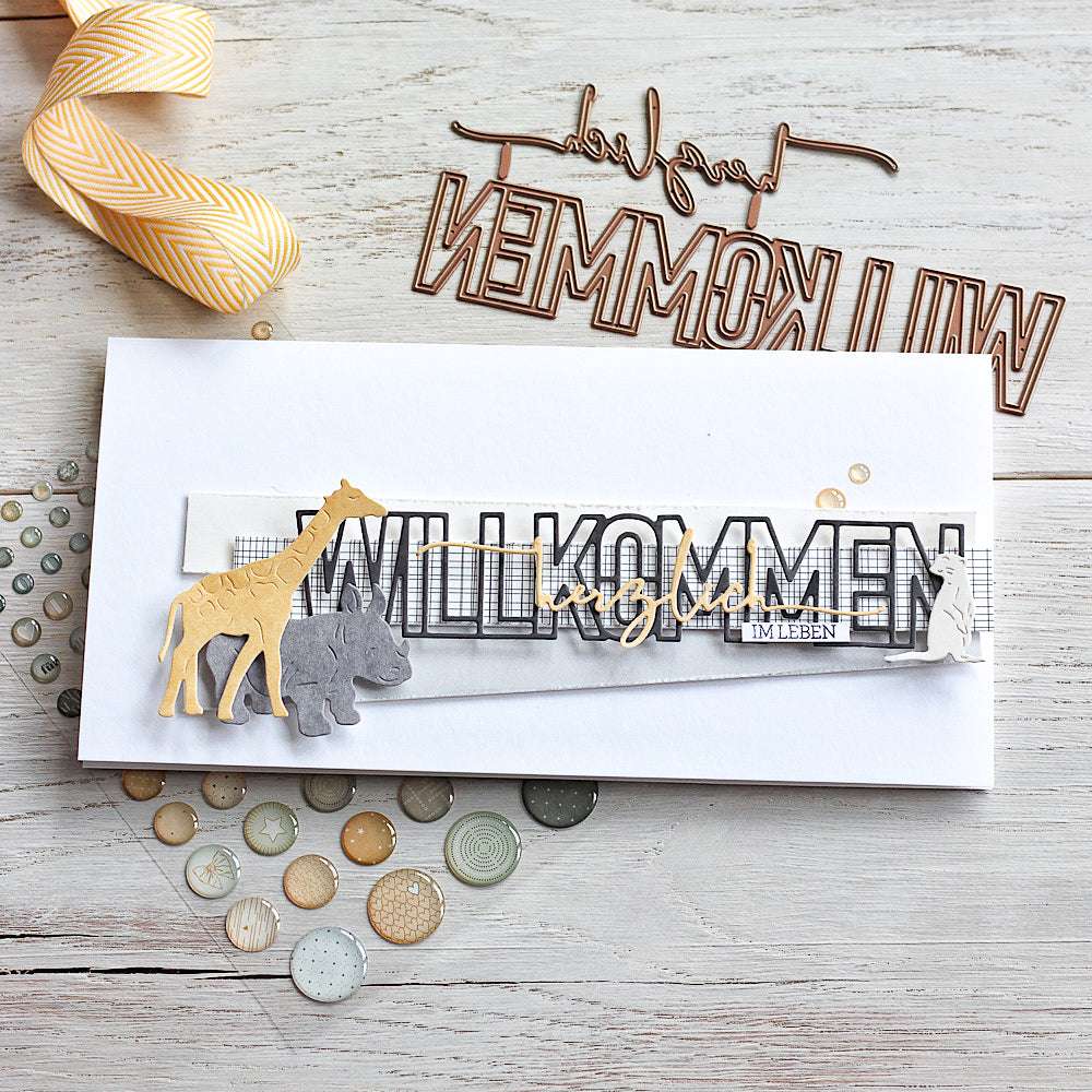 Handgemachte Karte mit dem Text "WILLKOMMEN", Tierausschnitten aus der Motivstanze Willkommen von Alexandra Renke, Schleife und dekorativen Knöpfen auf Holz. Ideal für Ihr nächstes Designpapiere-Projekt!.