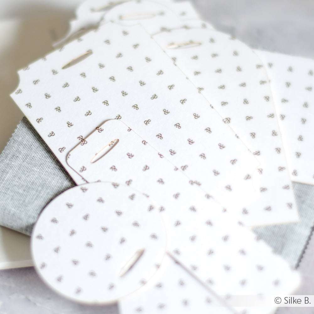 Alexandra Renke weiße Papieranhänger mit kleinen schwarzen Ampersand-Mustern, perfekt für Papierprojekte oder Buchbinderei, sind auf einer grauen Stoffoberfläche verstreut.