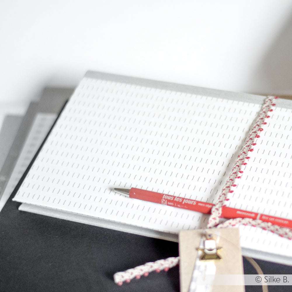 Ein roter Stift liegt auf einem weiß gemusterten Designpapier-Notizbuch von Alexandra Renke, das mit einer dekorativen Schnur und einem braunen Anhänger gebunden ist.