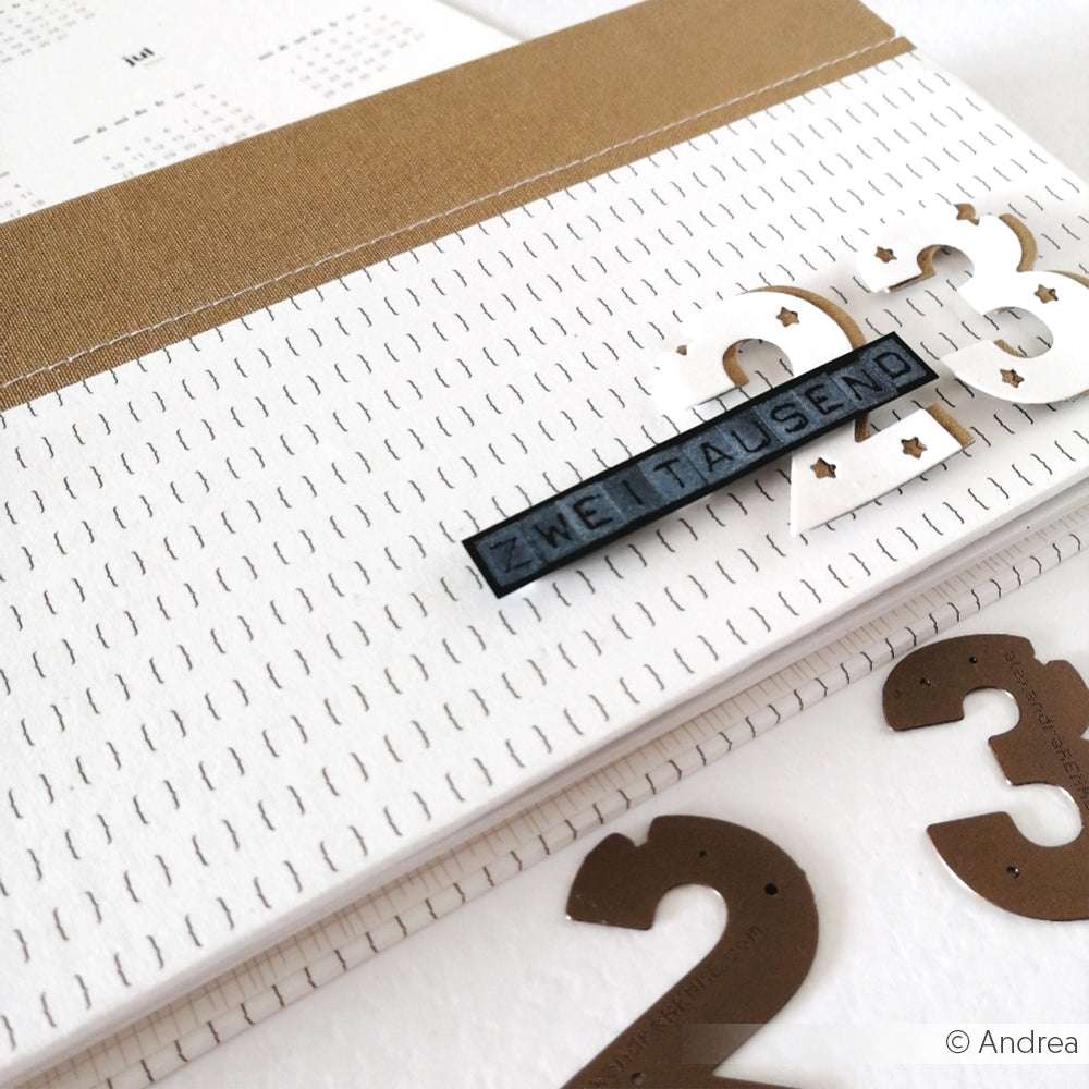 Nahaufnahme eines Alexandra Renke Kalenders mit "2023" und "ZWEITAUSEND" auf einem schwarzen Etikett, akzentuiert durch goldene Klammern-ideal für Papierprojekte oder kreative Designpapier-Inspirationen.