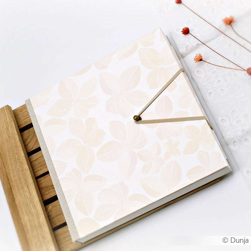 Ein floral gemustertes Scrapbook von Alexandra Renke, hergestellt mit Blüten Muster Pastellorange Designpapier und einem beigen Gummiverschluss, ruht auf einem Holztablett neben weißem Stoff und getrockneten Blumen. Ideal für Papierprojekte und Inspiration.