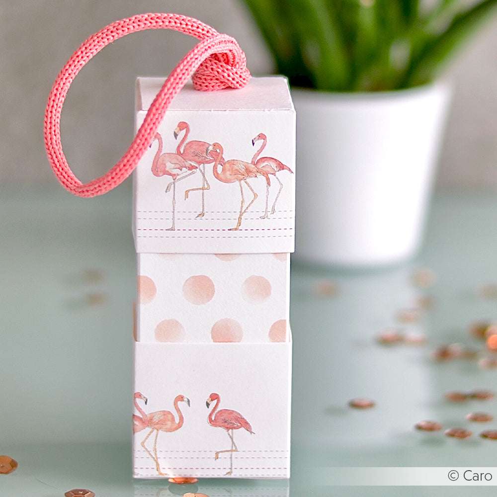 Auf einem Glastisch steht eine kleine Geschenkbox von Alexandra Renke aus Flamingo- und Polka-Dot-Designpapier, verziert mit einer rosa Kordel - ideal für Papierprojekte oder als Inspiration für Buchbinderei-Fans.