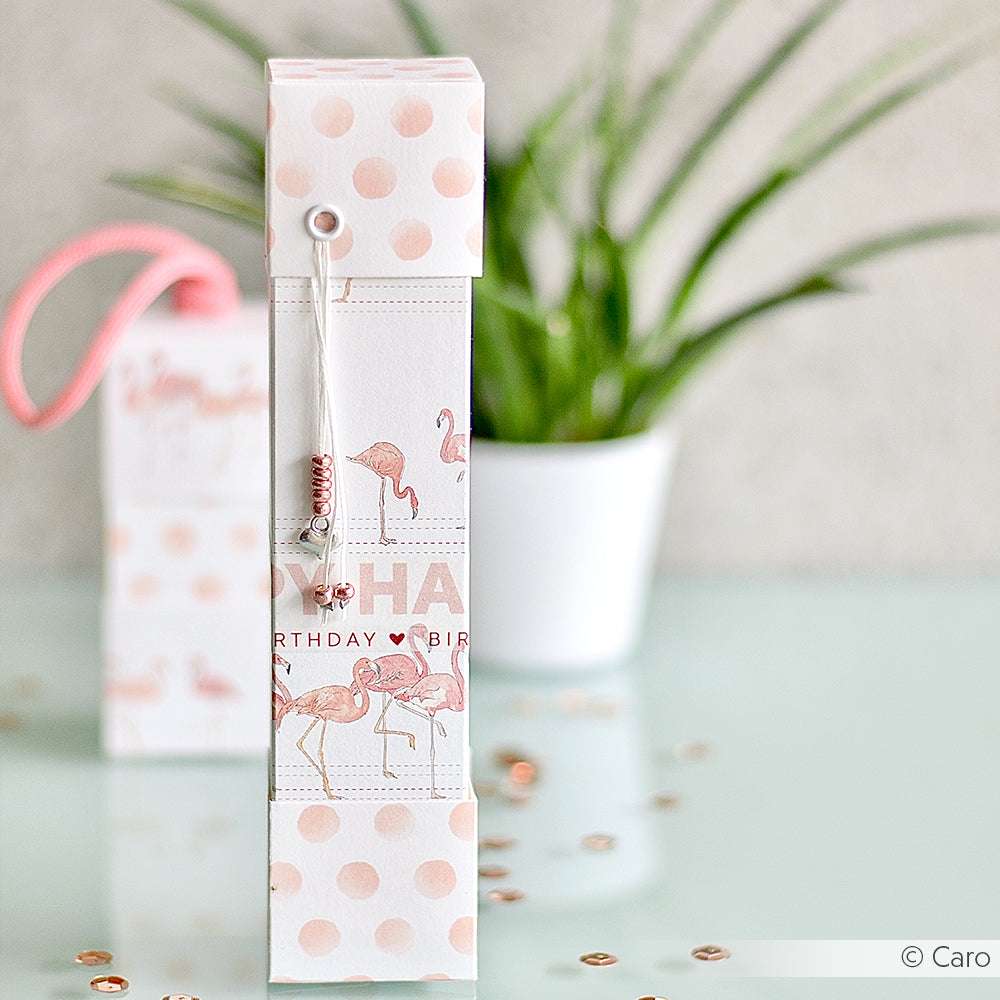 Die rosa-weiß gepunktete Geschenkbox mit Flamingo-Motiv von Alexandra Renke aus hochwertigem Designpapier setzt neben einer Pflanze und Pailletten einen reizvollen Akzent für Ihre Papierprojekte.