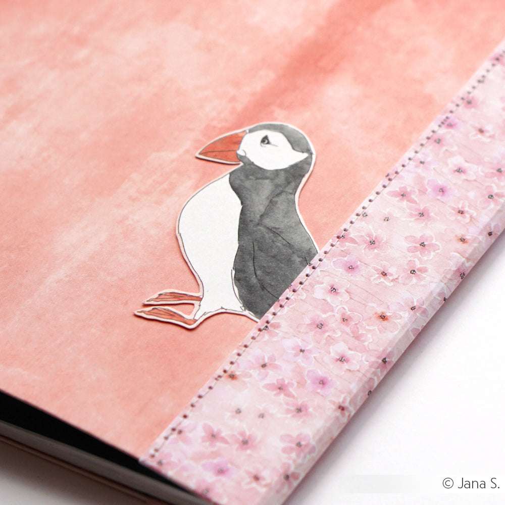 Ein Alexandra Renke Papageientaucher-Lesezeichen, inspiriert von Papageientauchern auf Aquarell, lugt aus einem rosafarbenen Notizbuch mit Blumenkranz auf einem pastellrosa Hintergrund hervor - ideal für Papierprojekte und kreative Gestaltungsideen.