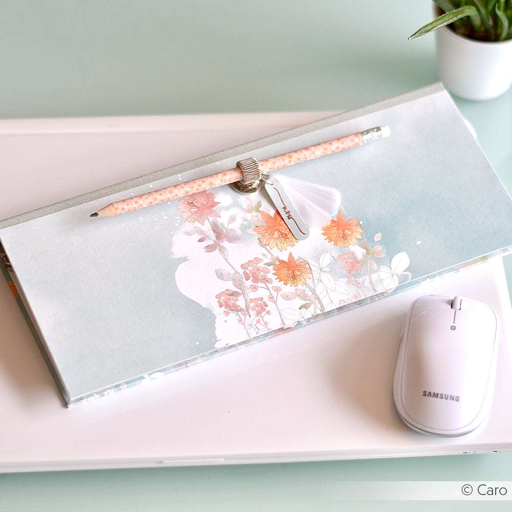 Ein geblümtes Notizbuch von Alexandra Renke, ein Designpapier-Stift mit Charme und eine weiße Samsung-Maus auf einem Glastisch neben einer Topfpflanze - ideal für kreative Projekte oder um den nächsten Geistesblitz zu notieren.