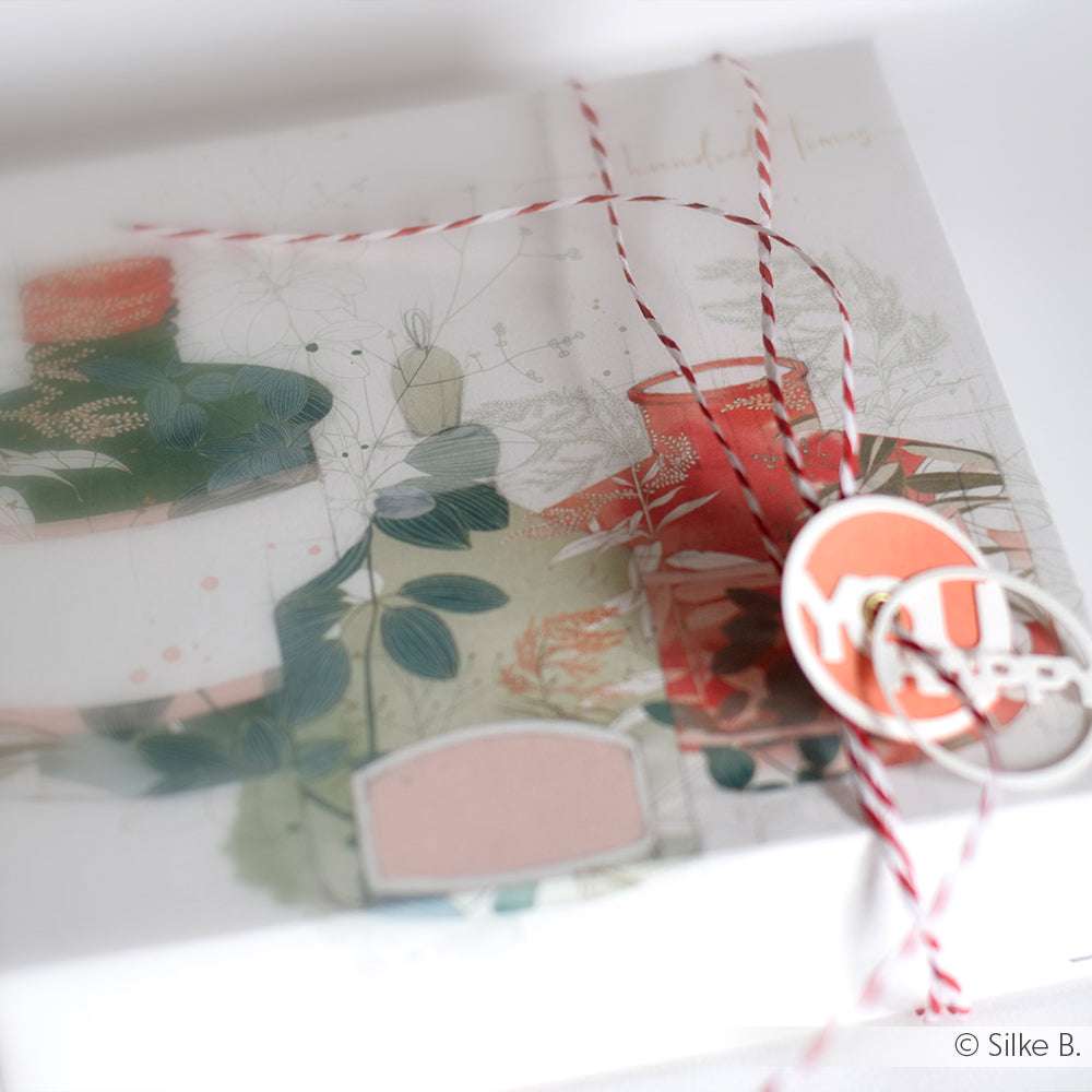 Als Geschenk verpackt in Alexandra Renke floralem Designpapier mit roter Kordel und dekorativen Anhängern, zart fokussiert - eine charmante Note für Ihre Papierprojekte.