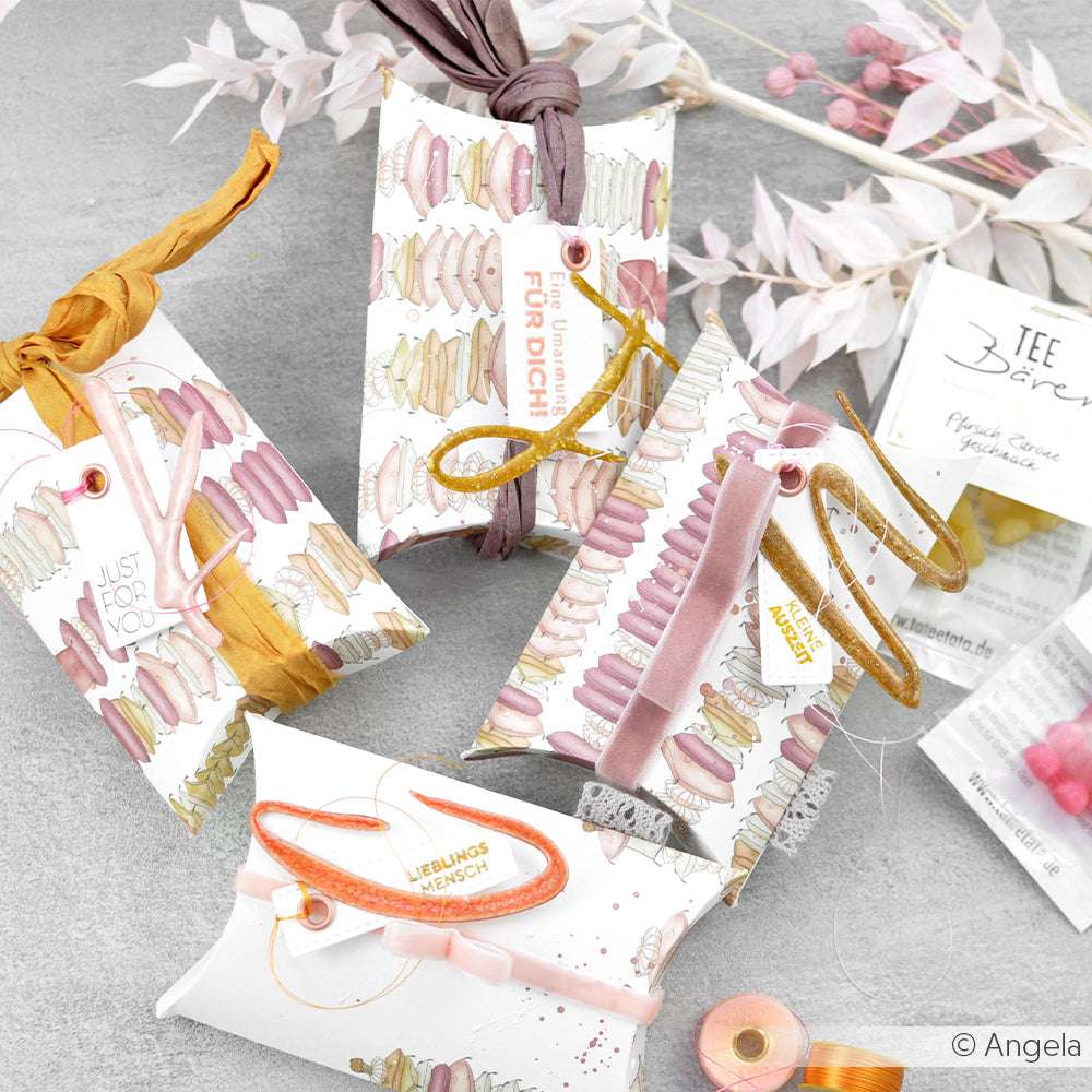 Auf einer hellgrauen Fläche sind Geschenkverpackungen mit pastellfarbenen Macaron-Motiven von Alexandra Renke arrangiert, die aus edlem Designpapier gefertigt und mit bunten Bändern verschnürt sind.