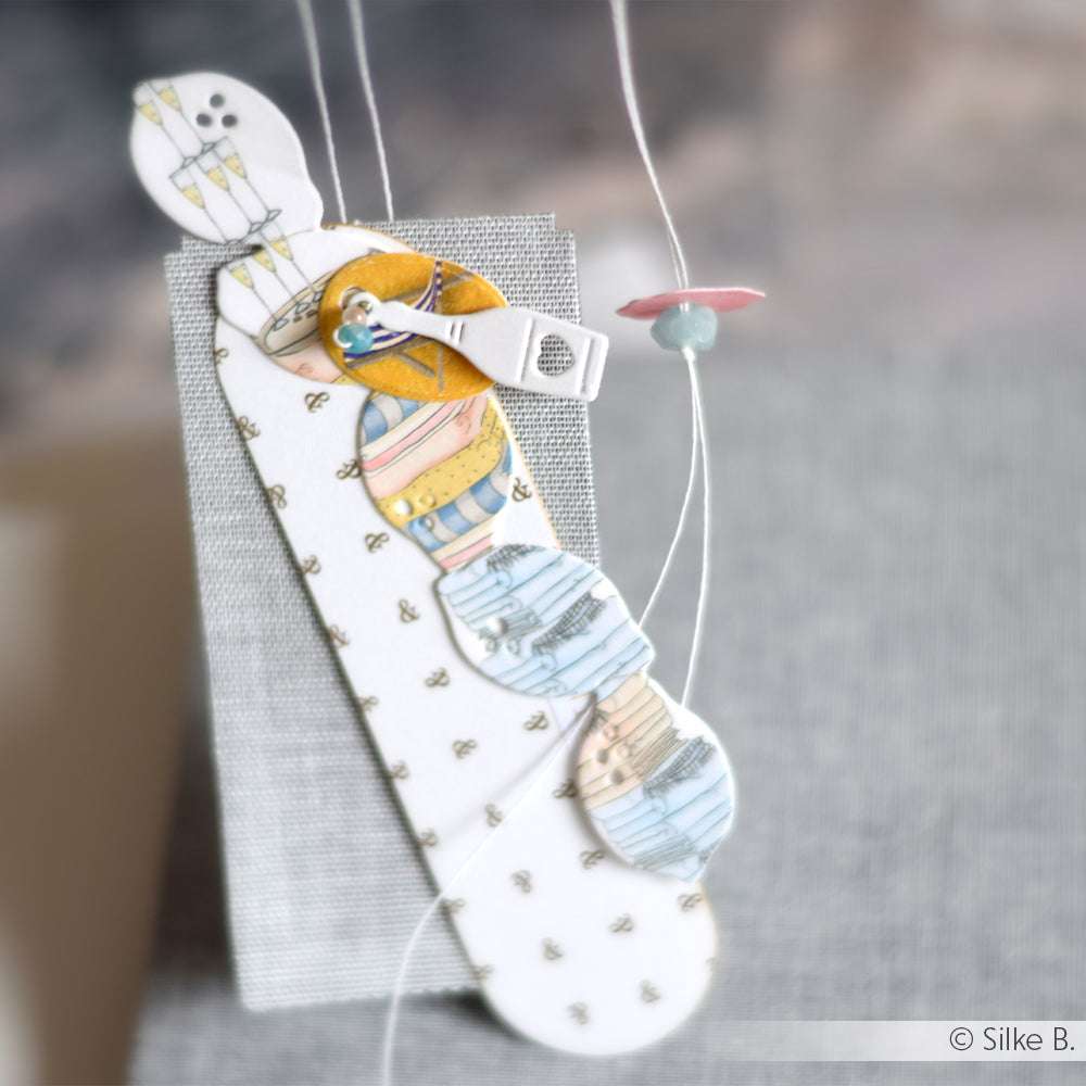 Handgeschöpftes Papier-Lesezeichen von Alexandra Renke aus Designpapier mit kreisförmigen Ausschnitten, Stoff- und Knopfdetails, elegant an Fäden hängend - ein auffälliger Akzent für Ihre Papierprojekte.