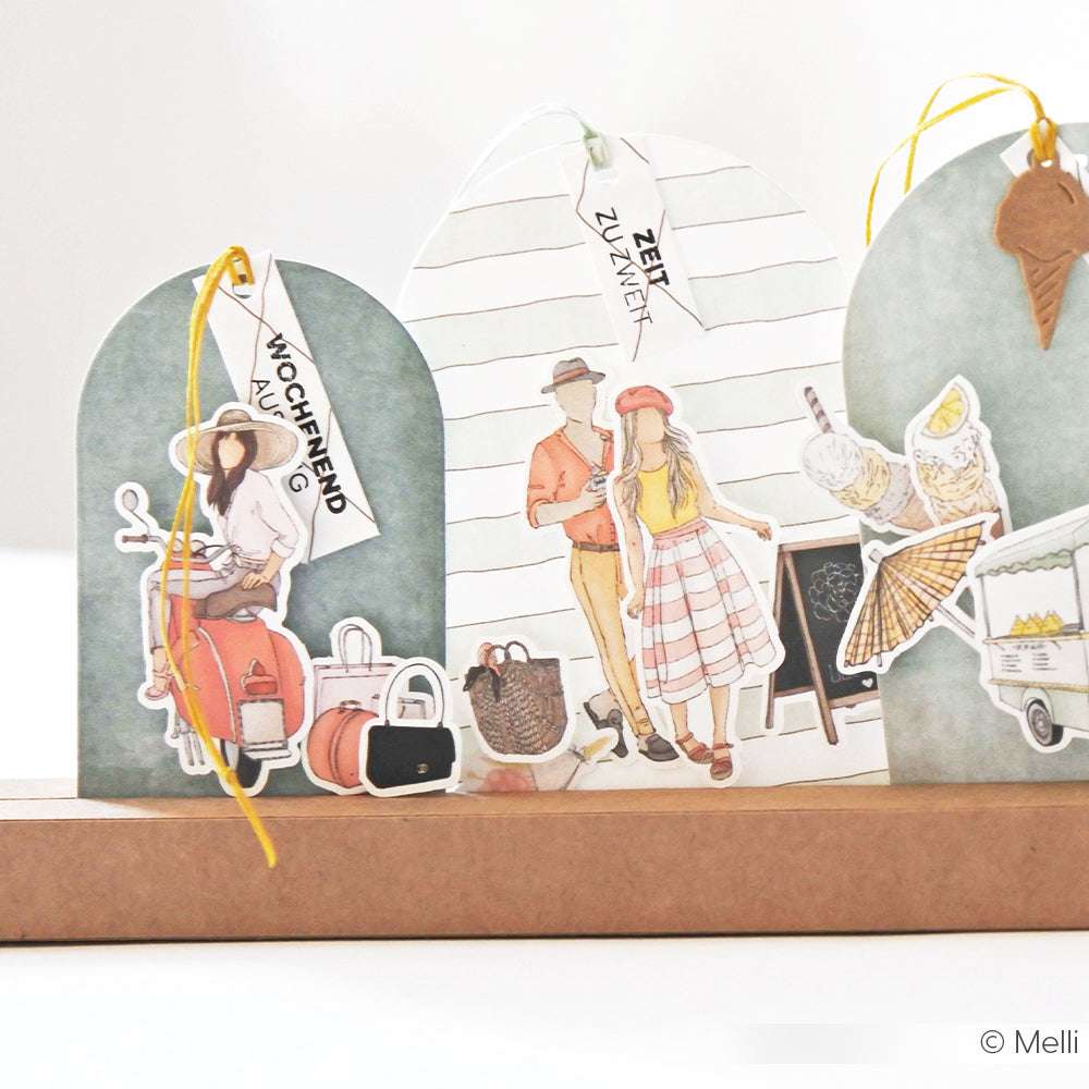 Illustrierte Scherenschnitte von stilvollen Reisenden und Marktartikeln, die mit dem exquisiten Alexandra Renke Designpapier gefertigt wurden, werden auf einem dekorativen Ständer präsentiert.