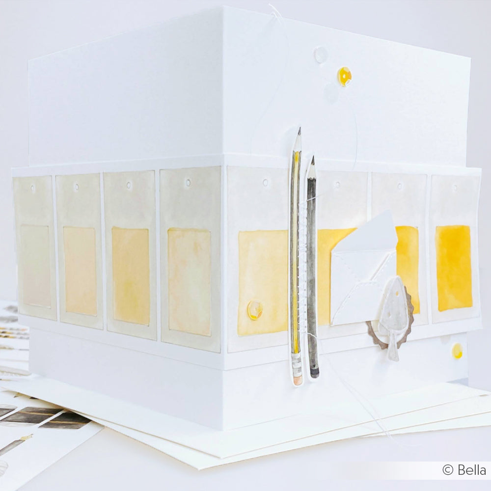 Eine weiße Alexandra Renke 3D Designpapier-Box mit gelben und grauen Akzenten, Bleistiften und einem kleinen Umschlag - ideal für Papierprojekte oder Buchbinderei.