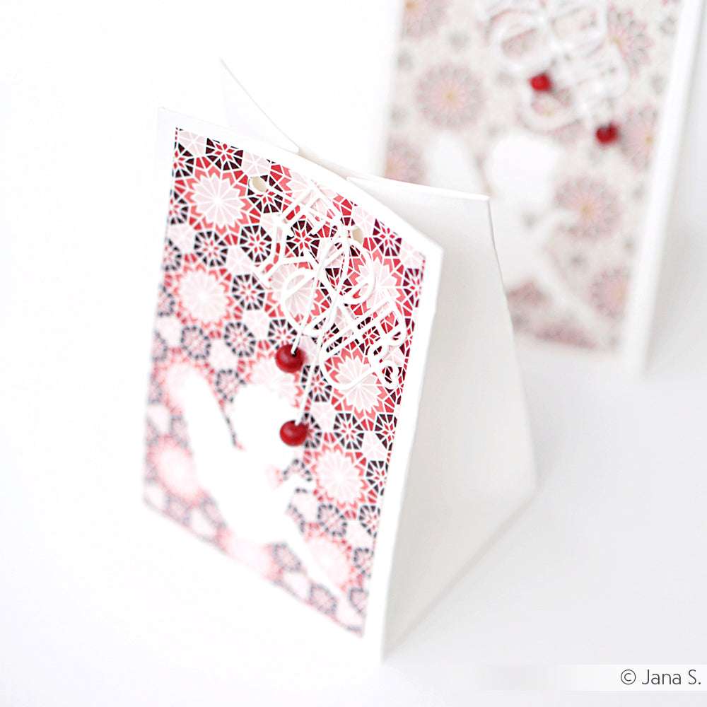 Eine Karte von Alexandra Renke mit einem weißen Vogel und roten Kirschen auf rotem, geometrischem Hintergrund, hergestellt aus Designpapier und aufrecht stehend.