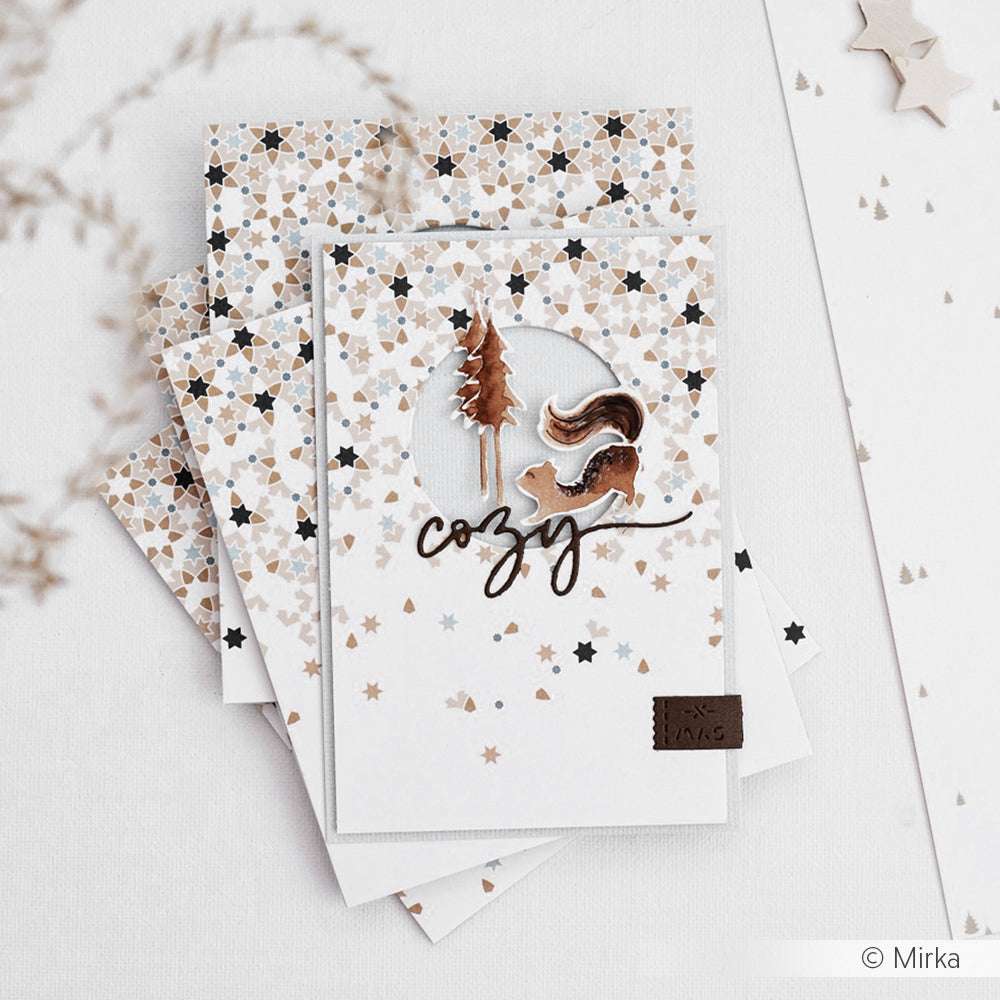 Handgemachte Karte von Alexandra Renke mit geometrischem Sternmuster, Baum, Eichhörnchen und gemütlichem Text in neutralen Tönen, gestapelt auf weiteren Papierprojekten.