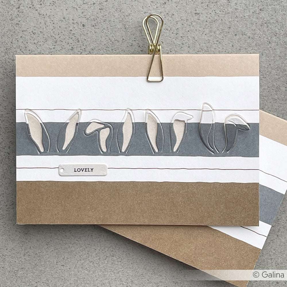 Eine handgefertigte Karte von Alexandra Renke mit Hasenohren auf neutralem, gestreiftem Designpapier und einem "LOVELY"-Etikett, das mit einer goldenen Klammer befestigt ist.