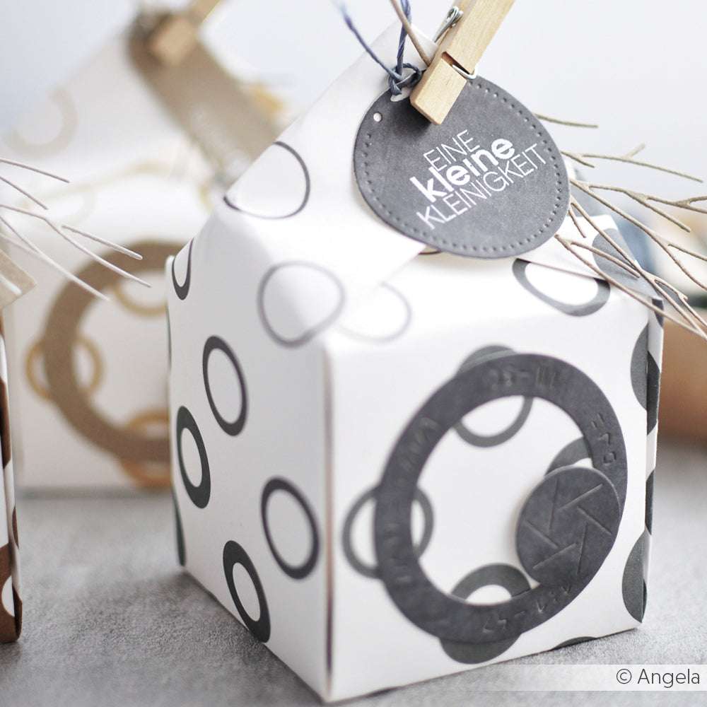 Eine kleine weiße Geschenkschachtel von Alexandra Renke aus Designpapier mit schwarzen Kreismustern, die mit einem runden Anhänger mit Clip versehen ist.
