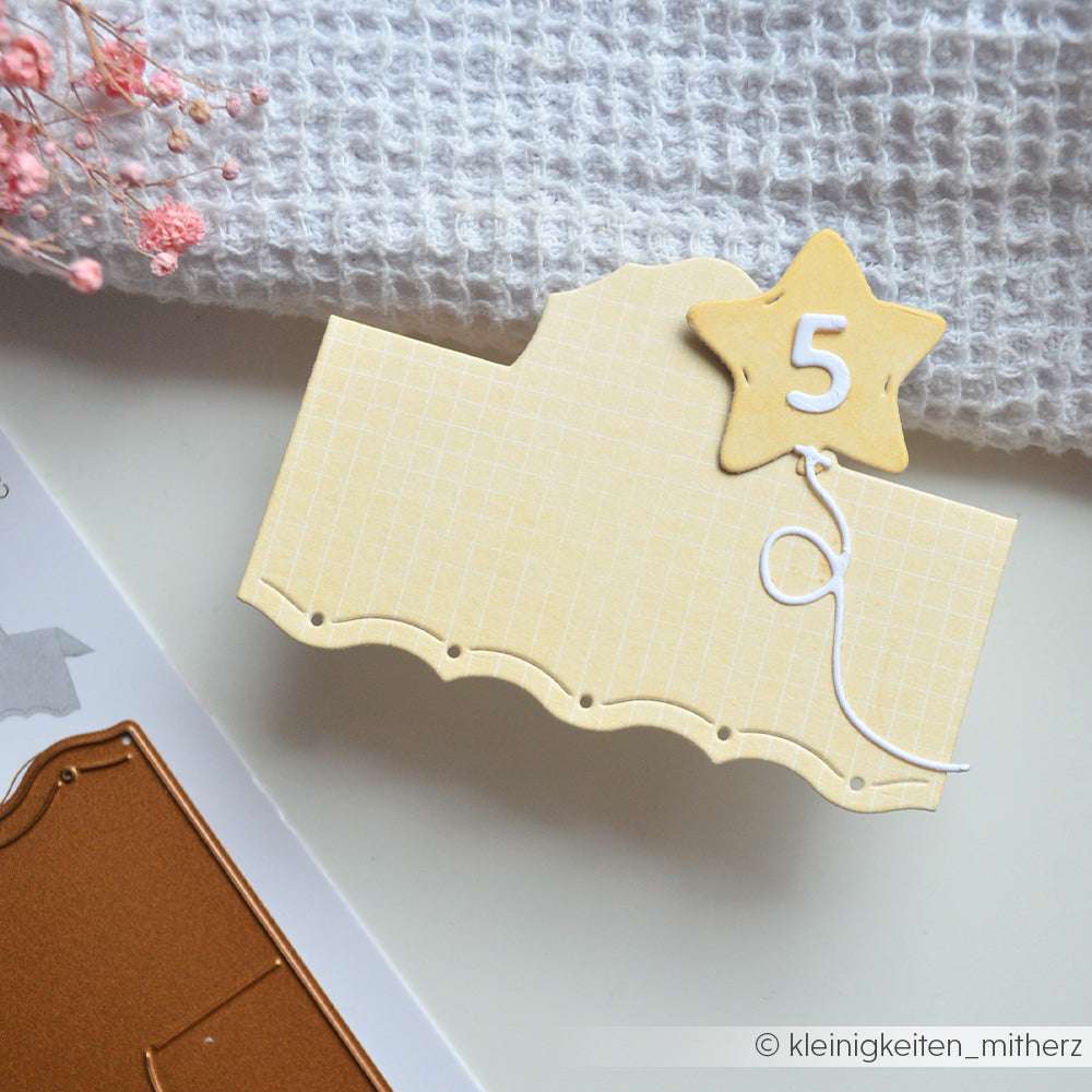 Eine gelbe Karte, die mit Alexandra Renke Mimi Zitronengelb Designpapier erstellt wurde, zeigt einen Sternenballon mit der Aufschrift 5, der auf einer weißen Fläche in der Nähe von strukturiertem Stoff und Bastelmaterial liegt.