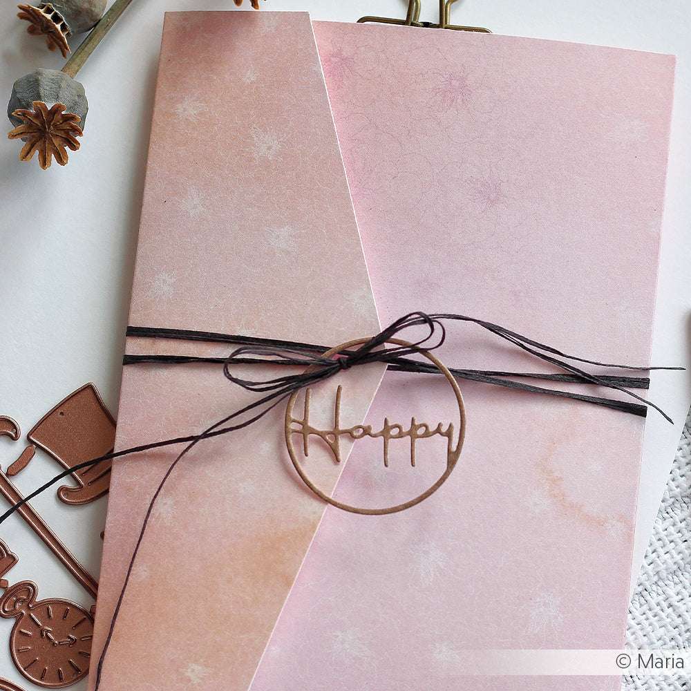 Eine rosafarbene Klappkarte von Alexandra Renke, gebunden mit schwarzer Kordel und einem goldenen Happy Charm, hergestellt mit Buchbinderei-Techniken und Designpapier Floral Apricot-Rose, wird auf einer weißen Fläche präsentiert.