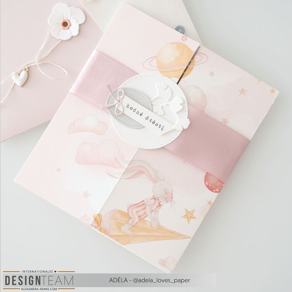 Rosa handgefertigte Karte aus Alexandra Renke-Papier mit einem Hasen auf Schultüte, der auf einer Sternschnuppe zwischen Planeten reitet und einem runden Anhänger mit der Aufschrift hodně štěstí.