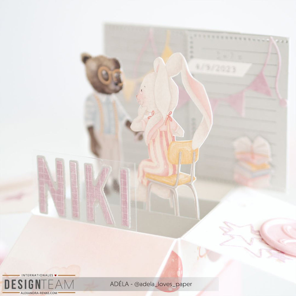 Pop-up-Karte von Alexandra Renke mit einem Hasen im Schlafanzug, einem Bären, pastellfarbenen Bastelpapier-Dekorationen und "NIKI" in rosa Buchstaben auf der Vorderseite.