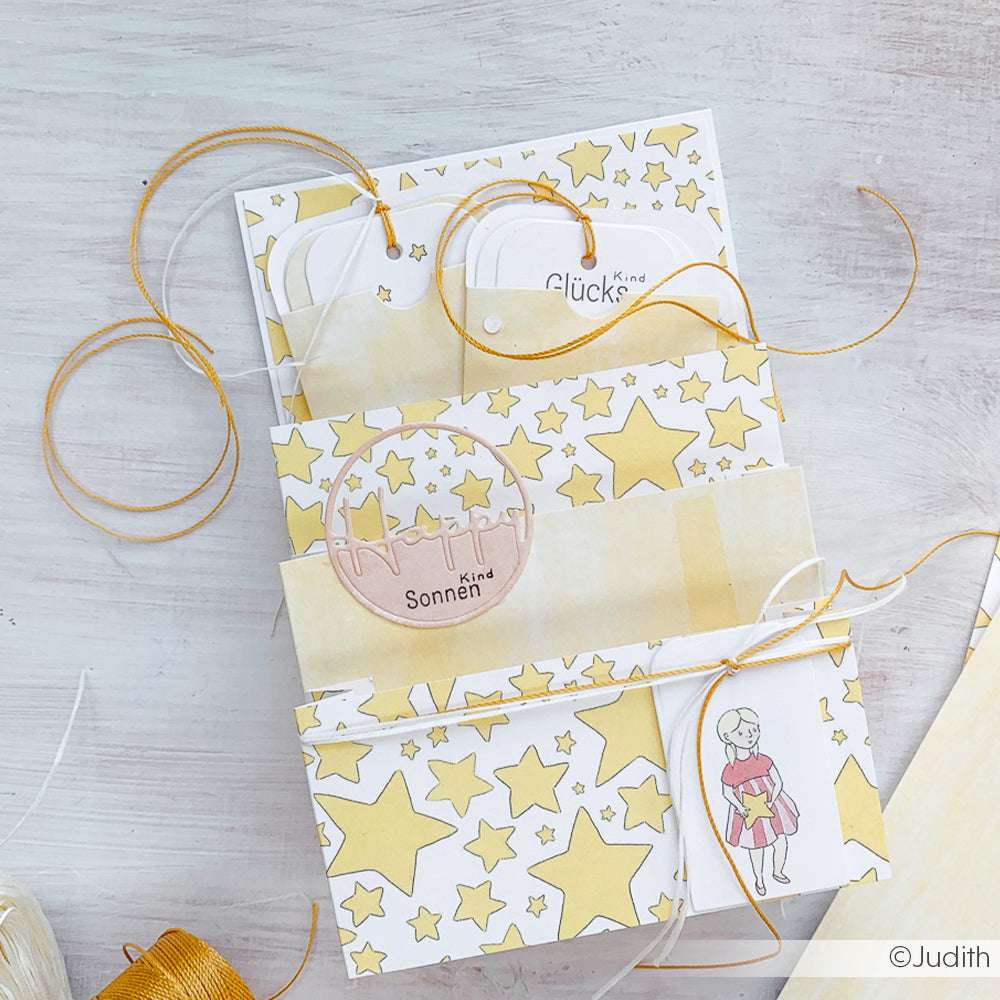 Handgefertigte Karte von Alexandra Renke, verziert mit gelben Sternen, orangefarbener Schnur und der Illustration einer Person, die ein Buch in der Hand hält.