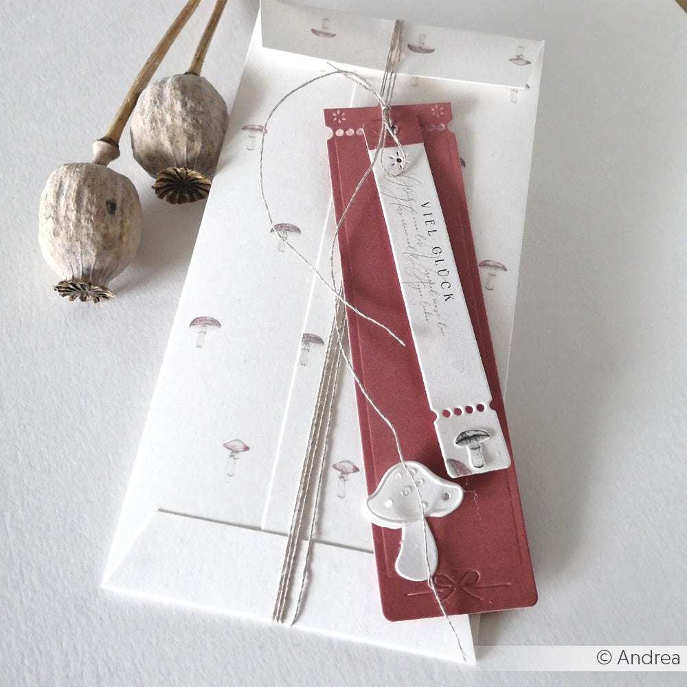 Weißer Umschlag mit Pilzmotiven, getrockneten Mohnschoten und einem roten Lesezeichen mit silbernem Pilzanhänger - perfekt für Papierprojekte oder Buchbinderei mit Ihrem Lieblingsdesignpapier von Alexandra Renke.