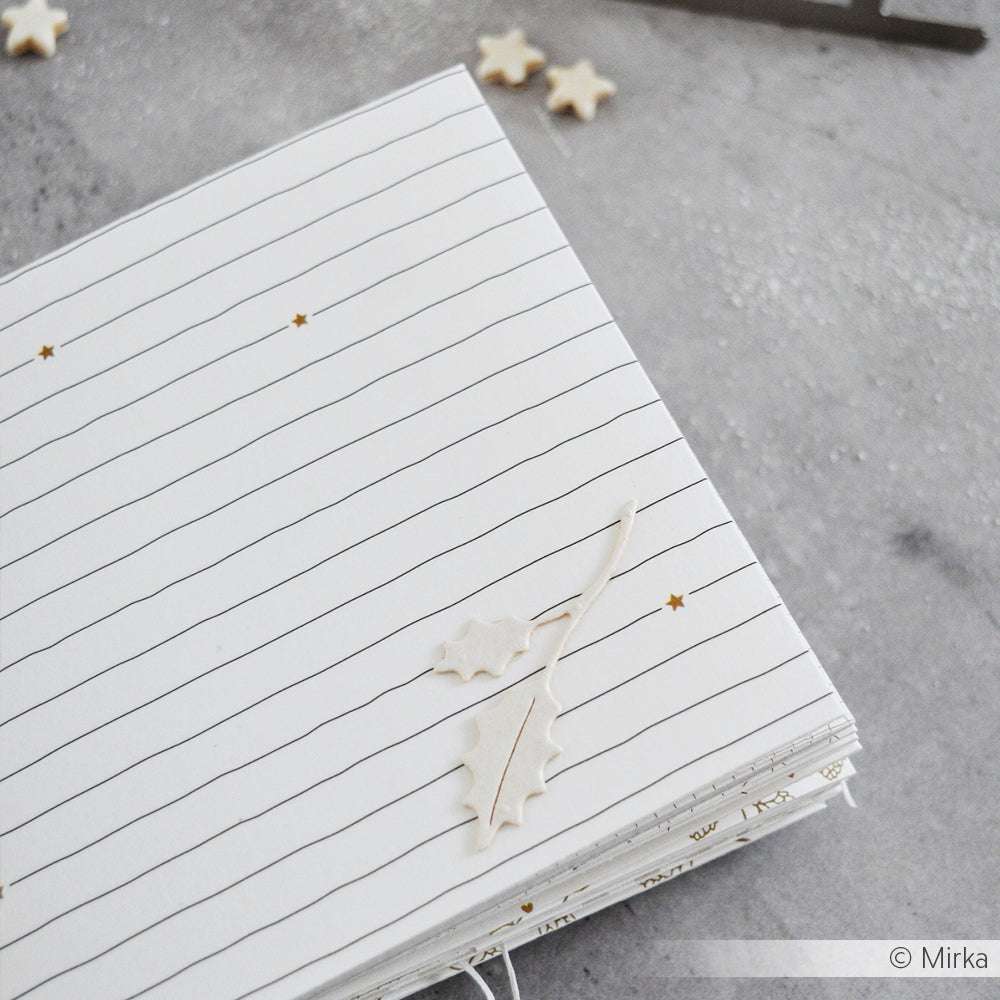 Alexandra Renke liniertes Notizbuch mit weißem Stechpalmenblatt-Dekor und kleinen Sternchen Linienmuster Gold Details, perfekt für Papierprojekte, dargestellt auf einer grauen Oberfläche.