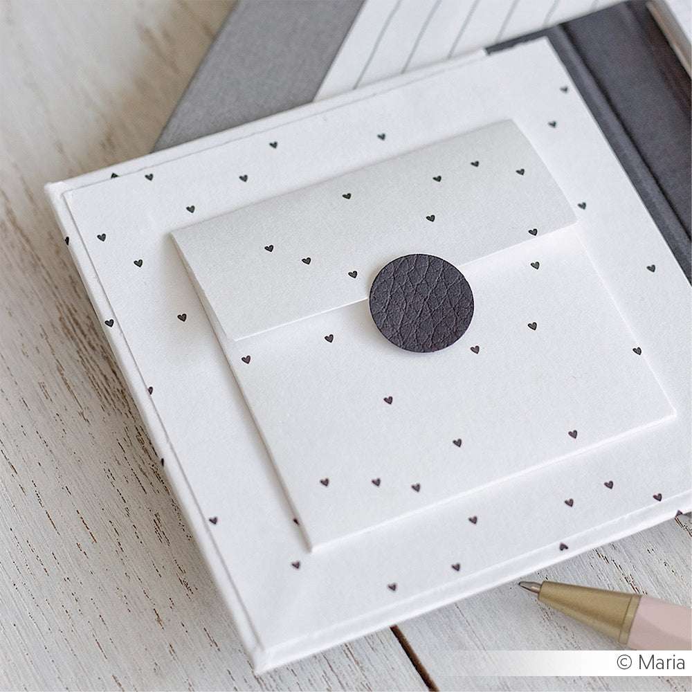 Weißer Umschlag mit kleinen schwarzen Herzen und einem runden Siegel von Alexandra Renke, dargestellt auf weißem Holz - ideal für Grußkarten oder Papierprojekte mit Designpapier Herzchenregen Schwarz.
