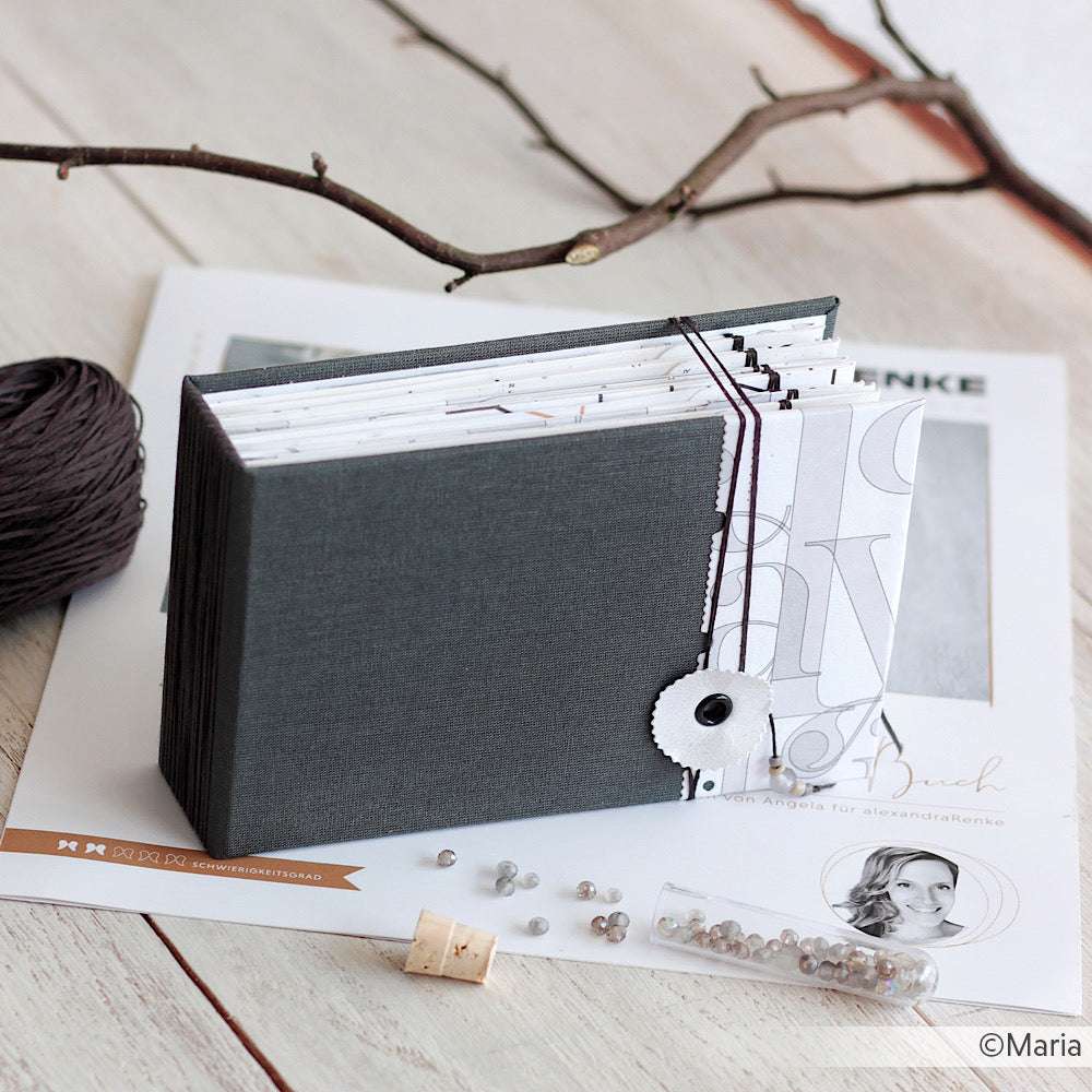 Das handgefertigte graue Scrapbook von Alexandra Renke mit Schnur und Knopfverschluss eignet sich perfekt für kreative Projekte oder einzigartige Karten, die auf einer weißen Fläche mit Bastelmaterial gezeigt werden.