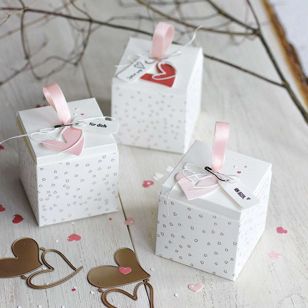 Drei weiße Geschenkschachteln mit rosa Schleifen und Herzanhängern, umgeben von Herzkonfetti, goldenen Herzklammern und eleganten Alexandra Renke Papierakzenten für eine stilvolle Präsentation.