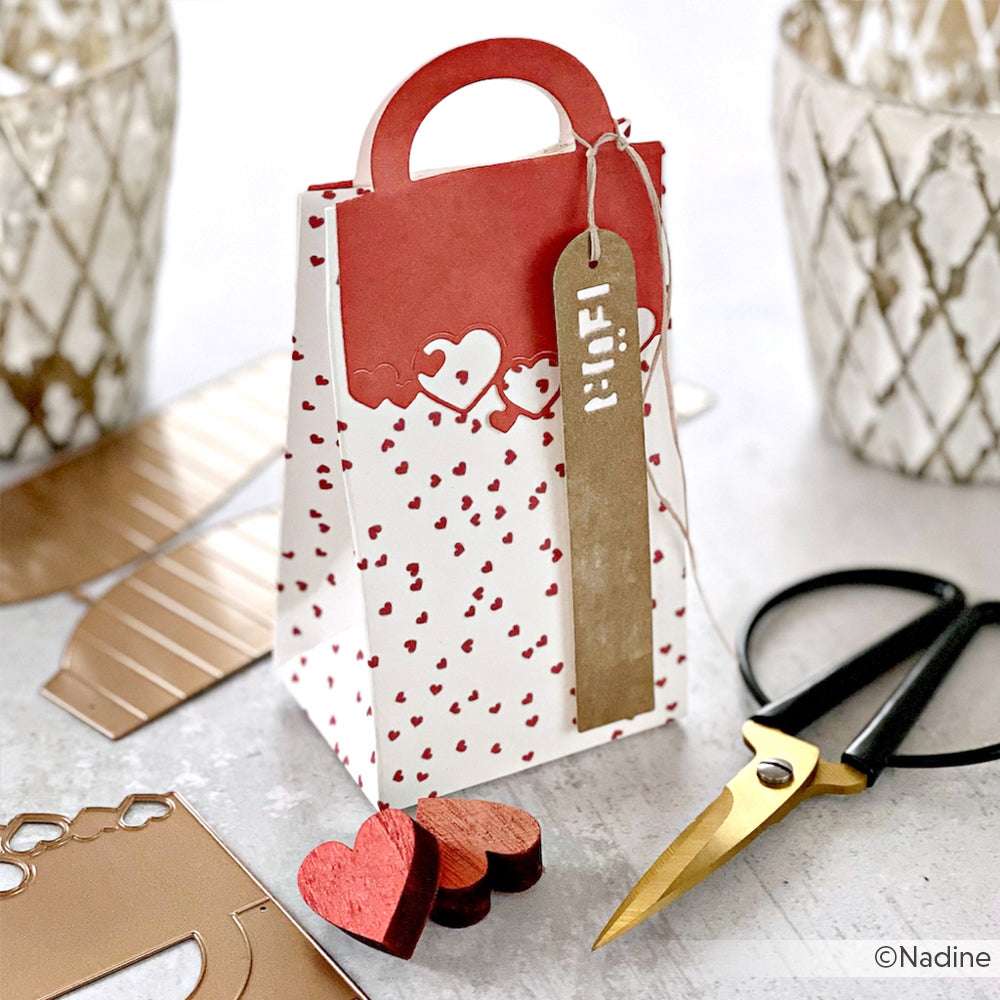 Eine Geschenktüte mit Herzmotiv von Alexandra Renke mit Anhänger, Schere, Designpapier und Bastelmaterial - ideal für Papierprojekte mit Herzgestöber Premiumrot - auf einer weißen Fläche.