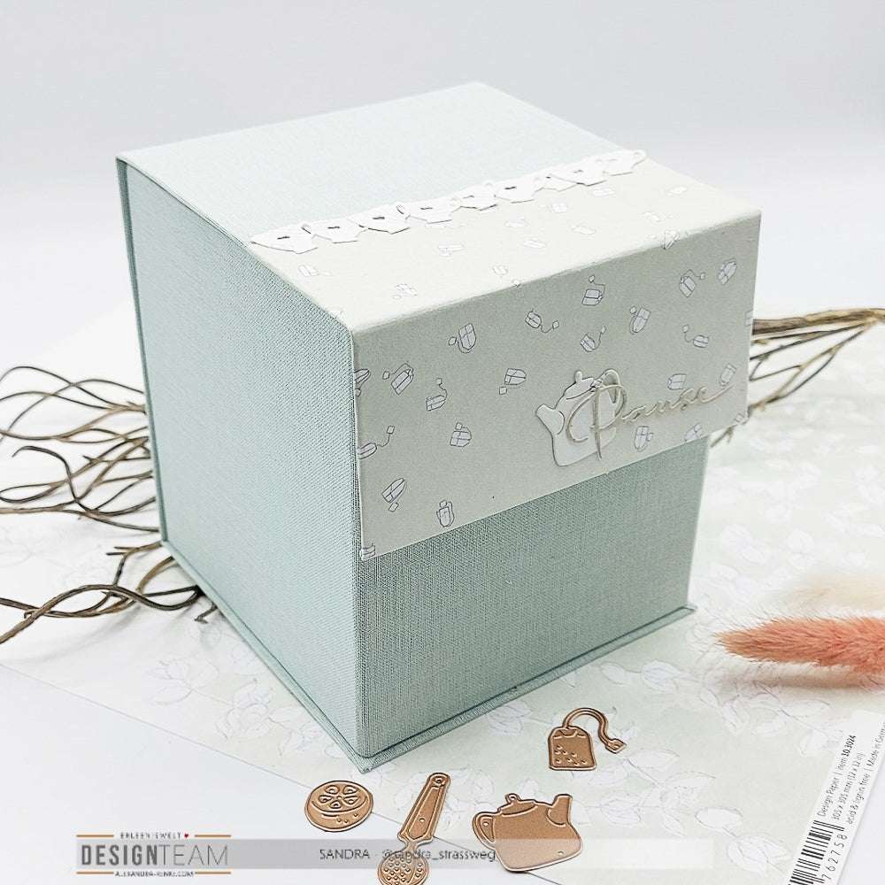 Eine hellblaue Alexandra Renke Geschenkbox mit einem gemusterten Deckel, inspiriert von Teekannen- und Tassenmotiven aus der Teewelt, ruht auf einer weißen Oberfläche.